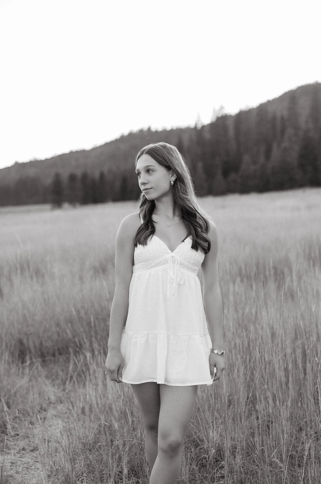 A young woman standing in a grassy field with hills and trees in the background, wearing a sleeveless white dress and looking to her left.