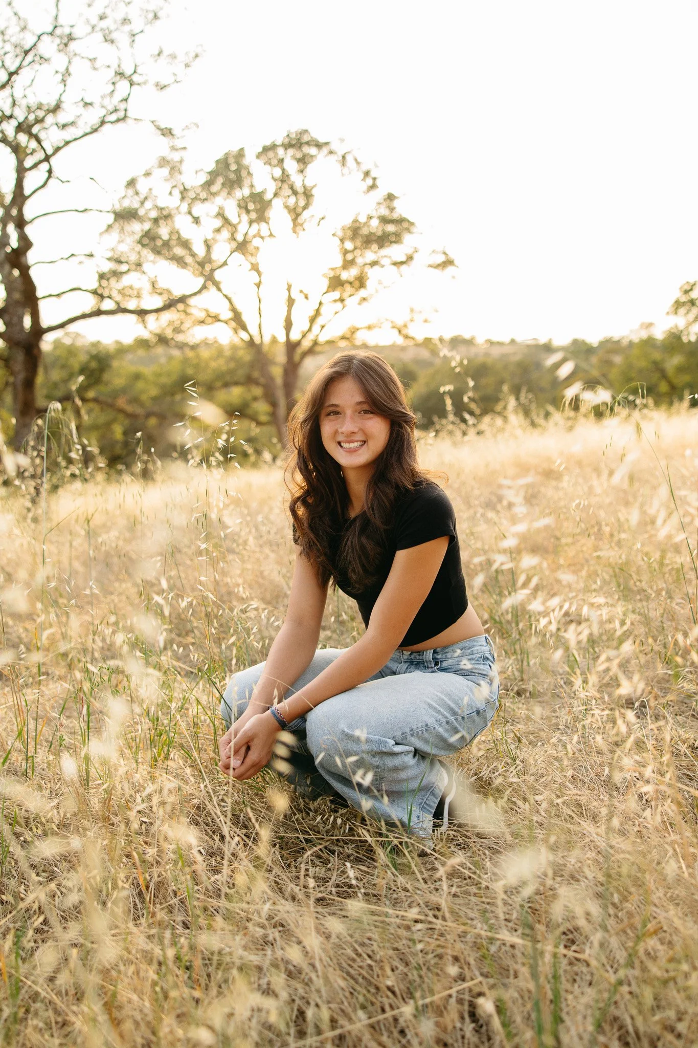 A young woman with long brown hair sitting in a field of tall grass during sunset, smiling at the camera, wearing a black t-shirt and light blue jeans.
