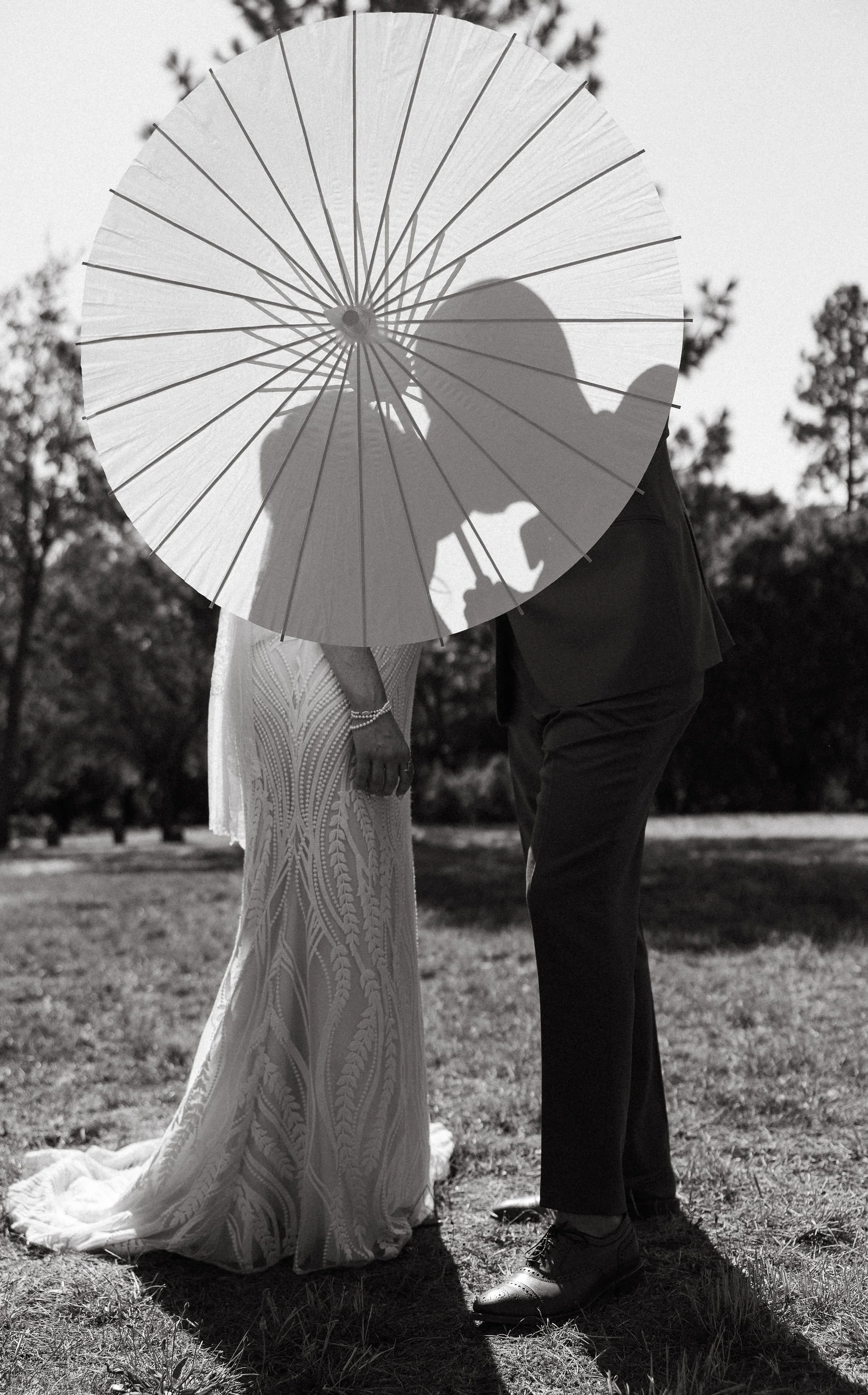 A black and white photo of a couple outdoors, with their faces hidden behind a large parasol. The woman is in a long, detailed dress, and the man is in dark pants and a jacket, leaning toward her, sharing an intimate moment.