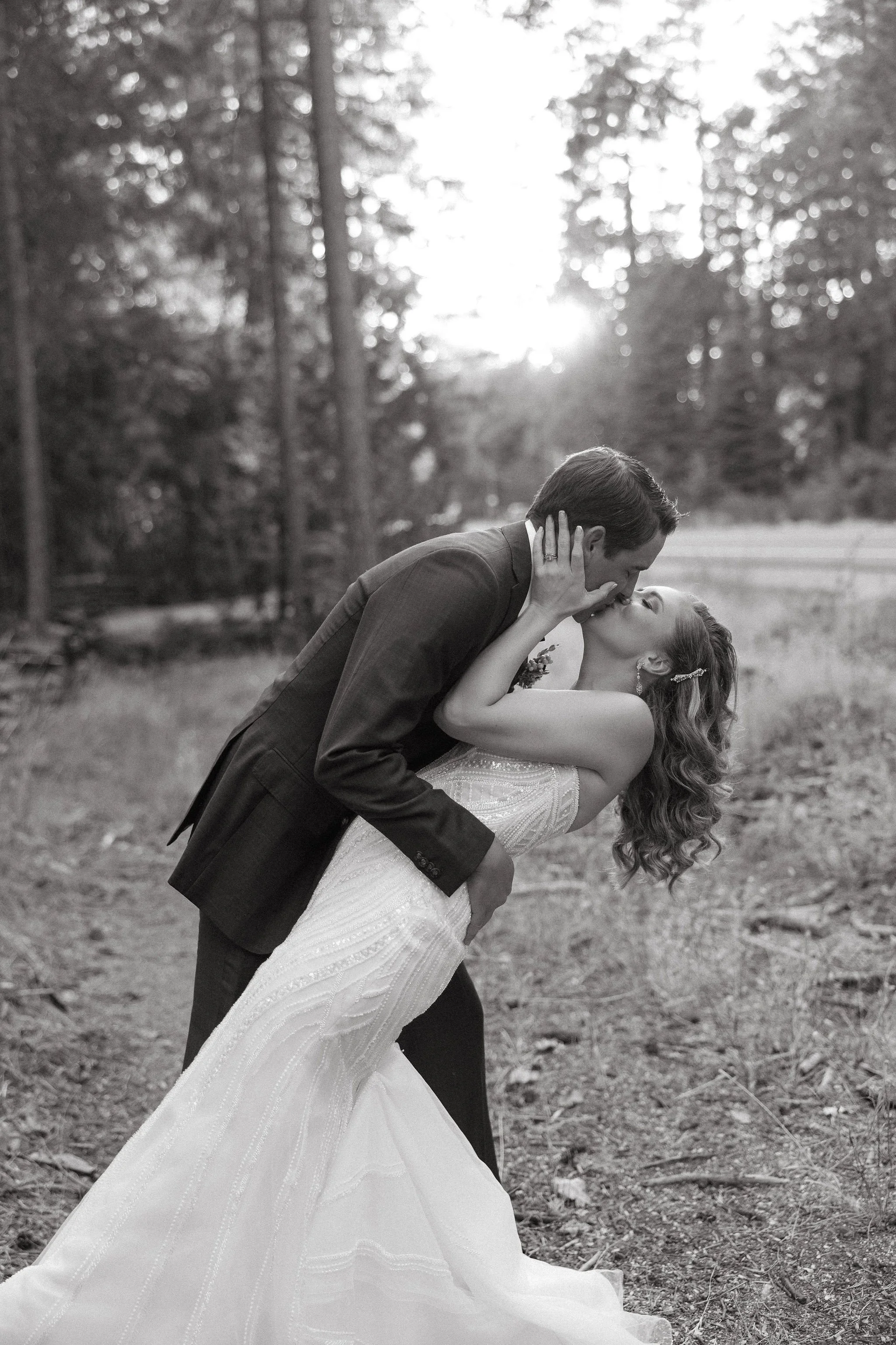 A black and white photo of a couple in wedding attire sharing a kiss outdoors, with trees and sunlight in the background.