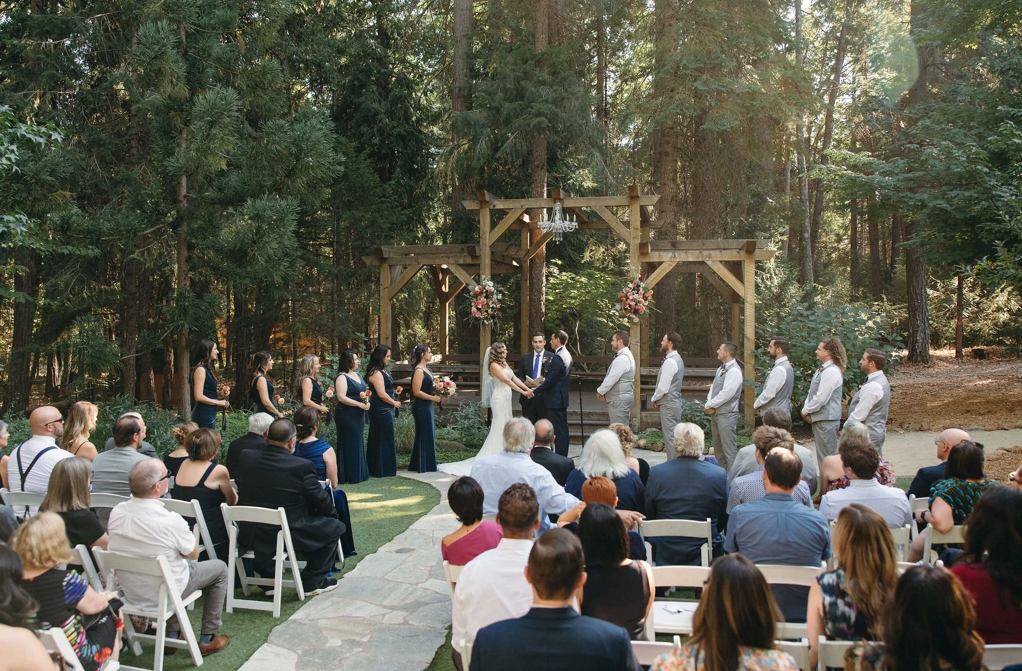Outdoor wedding ceremony taking place in a forest setting with a wooden arch decorated with flowers, where a bride and groom are exchanging vows. Bridesmaids and groomsmen stand on either side, and seated guests watch the ceremony.