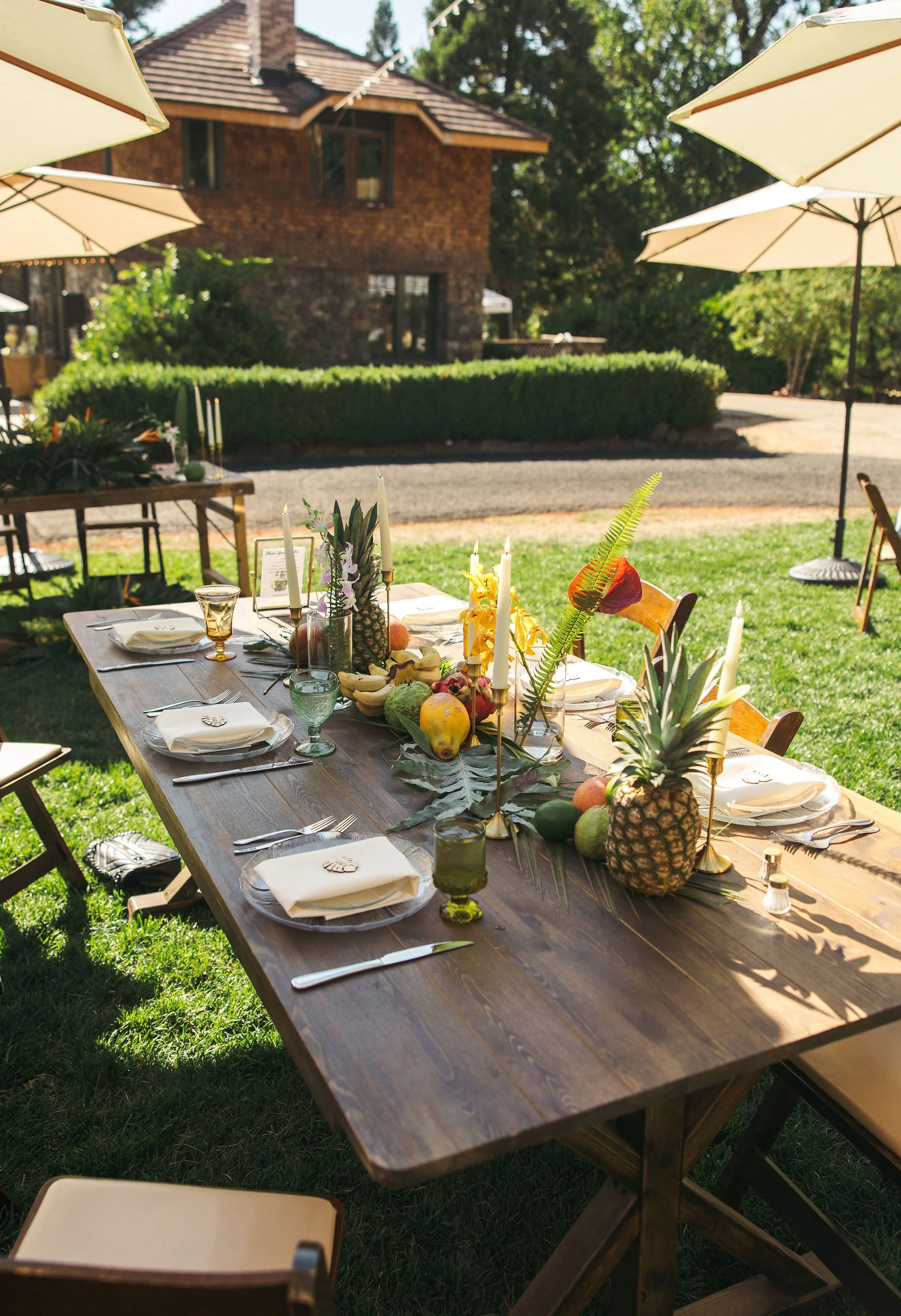 Outdoor dining table set with centerpiece of fruit, candles, and greenery under umbrellas on a sunny day.