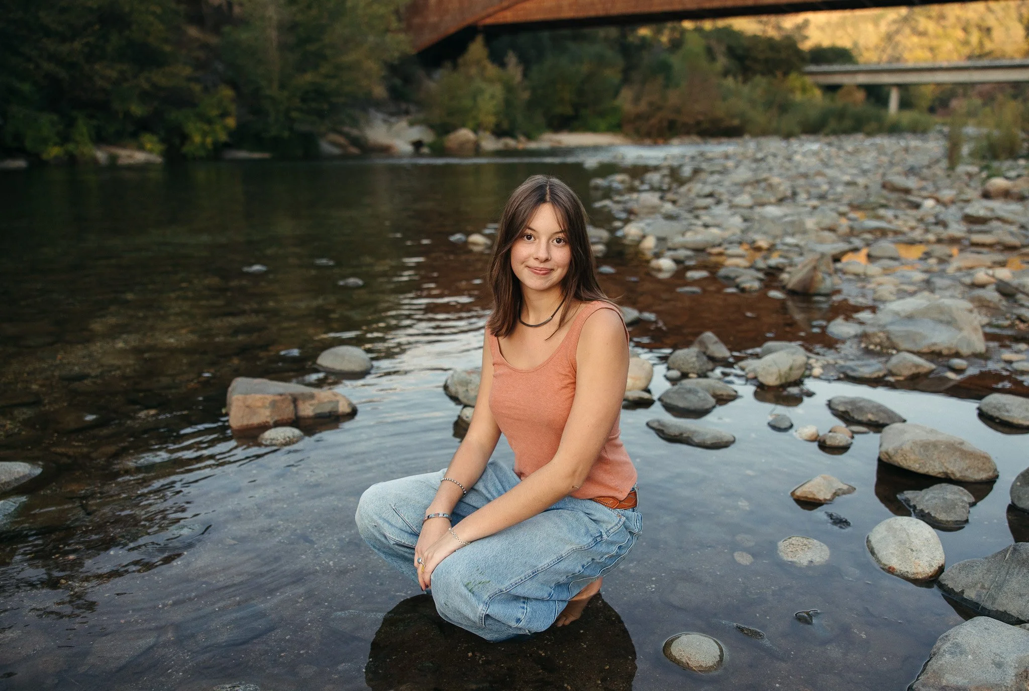 A young woman with brown hair sits in a shallow river with rocks, smiling at the camera. She is wearing a sleeveless top and jeans, with trees and a bridge in the background during what appears to be late afternoon or early evening.
