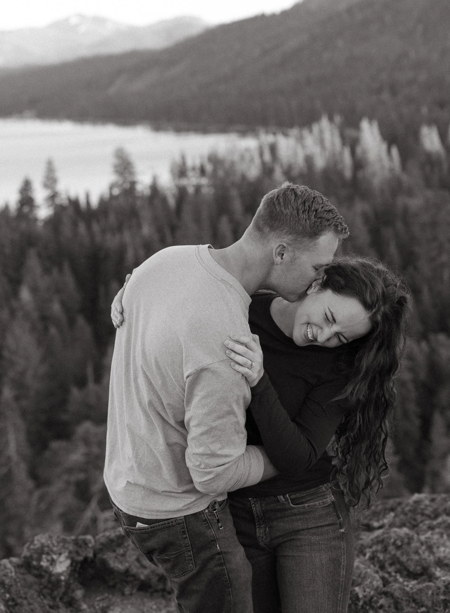A black and white photo of a smiling couple close together outdoors, with his head leaning against hers, in a natural setting with mountains and trees in the background.