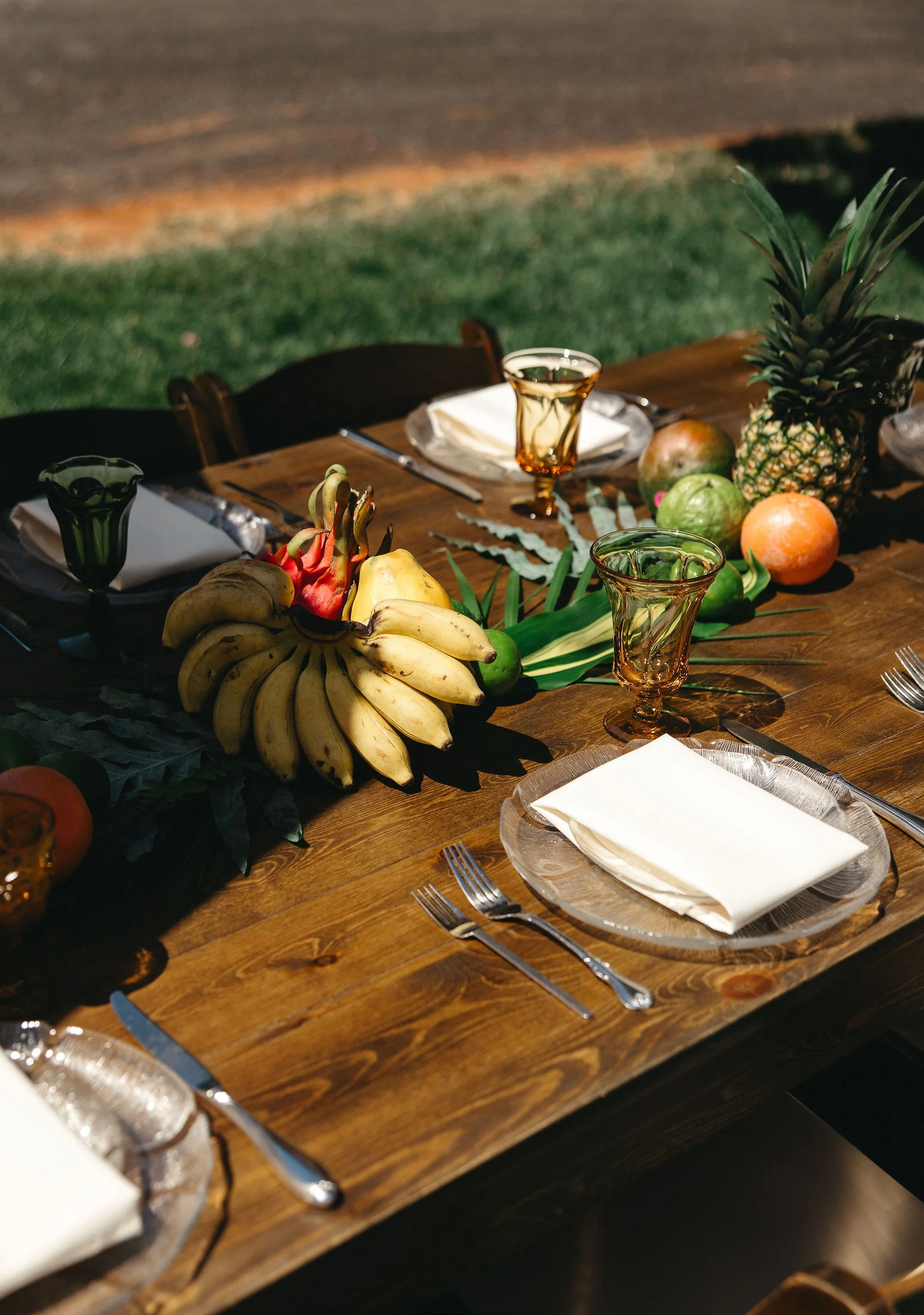 Outdoor dining table with tropical fruits, including bananas, pineapple, and other fruits, decorated with greenery, set with glassware, silverware, and white napkins.