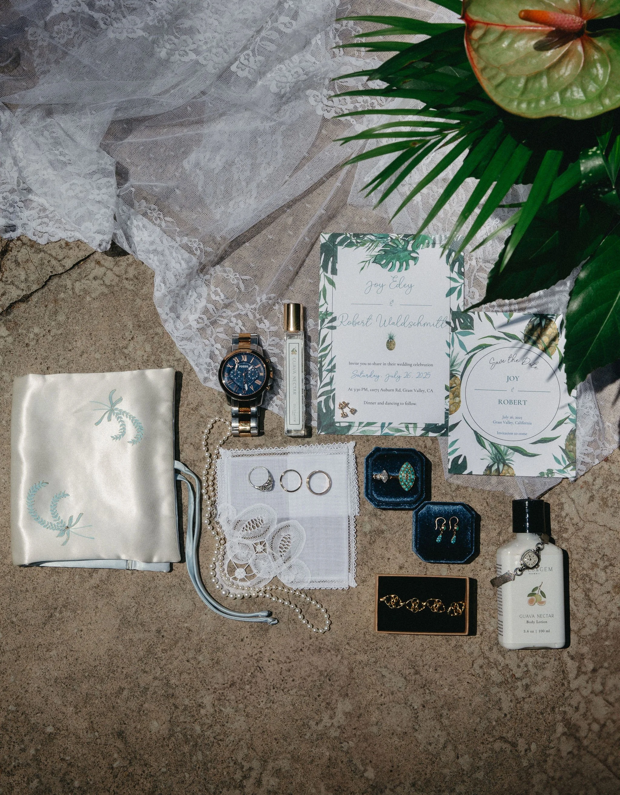 Wedding invitation, jewelry, watch, perfume, skincare, and other accessories on a sandy surface with white lace and tropical foliage.