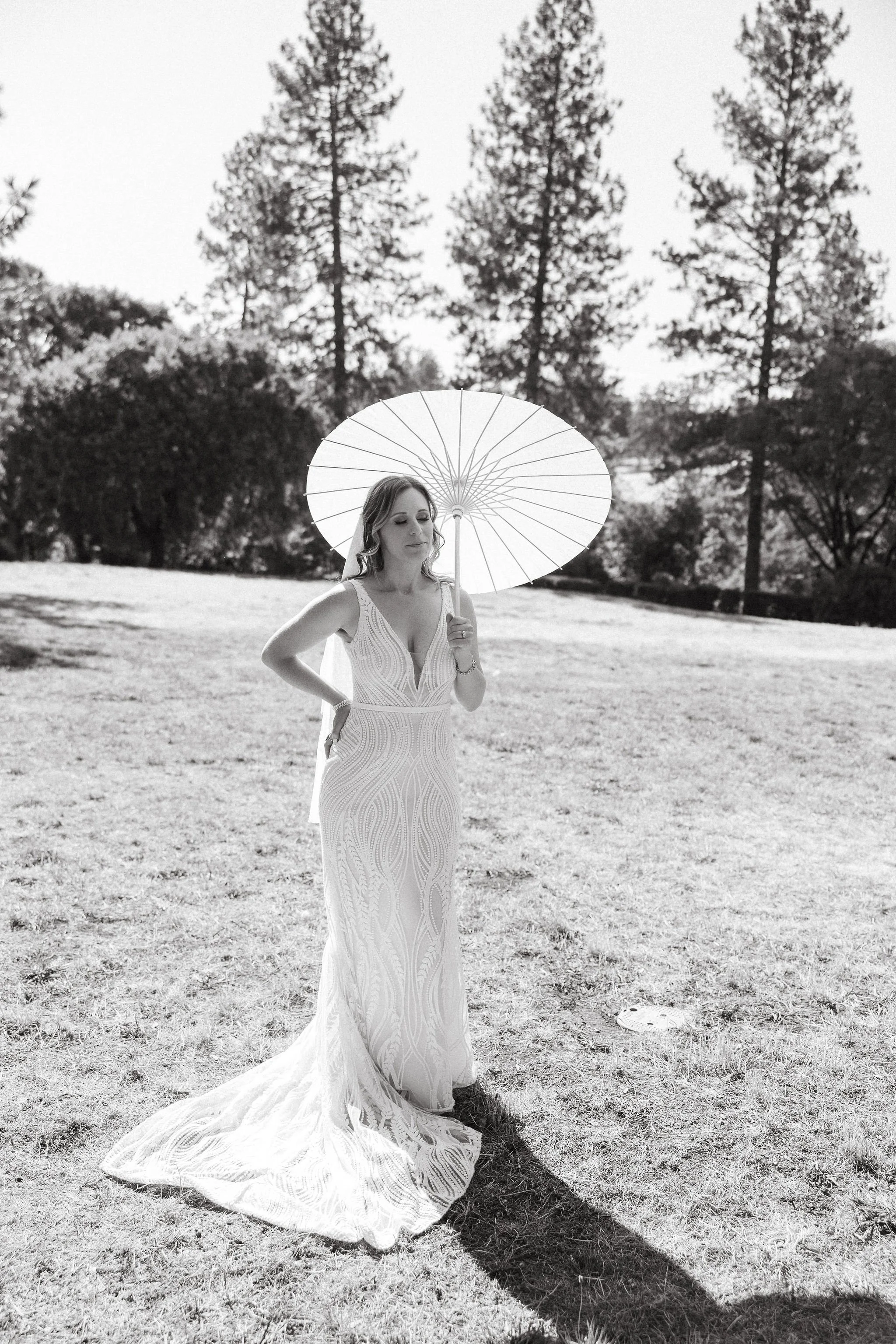 A woman in a white dress holding a parasol standing outdoors on a grassy area with trees in the background.