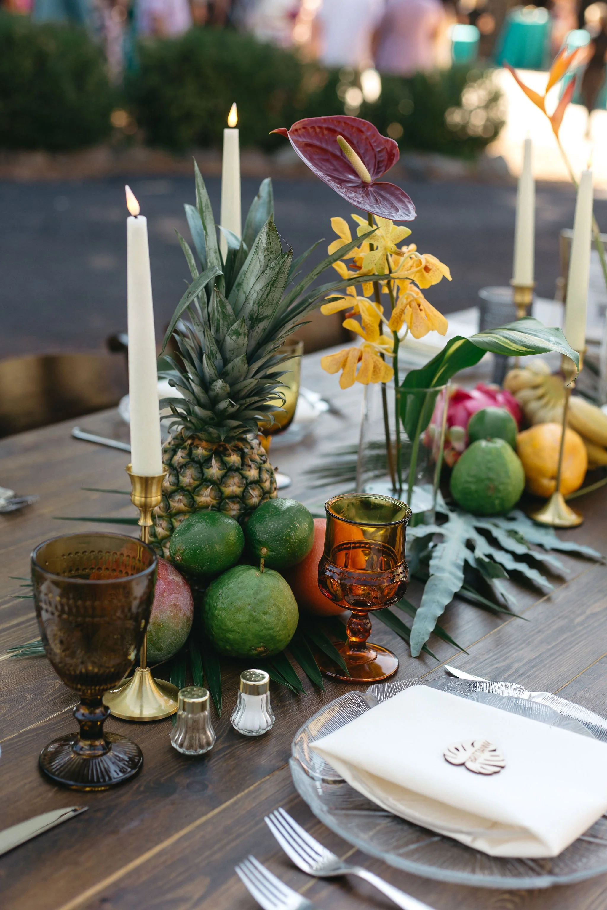 A rustic outdoor dining table decorated with tropical fruits including pineapple, limes, and avocados, along with colorful orchids, tall candles, and glassware.
