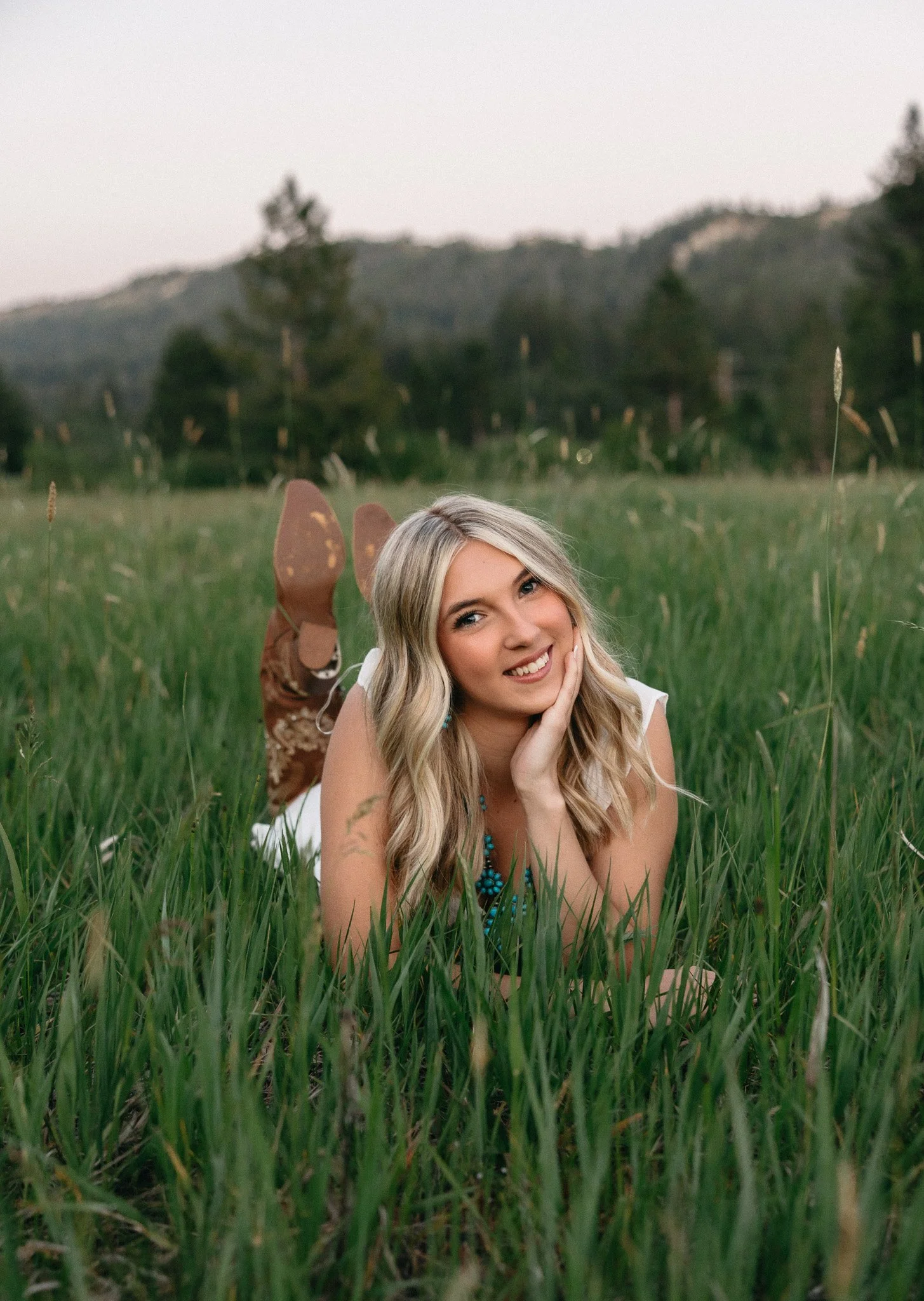 A young woman with long blonde hair lying on her stomach in a grassy field, smiling and resting her chin on her hand, with trees and hills in the background during dusk.