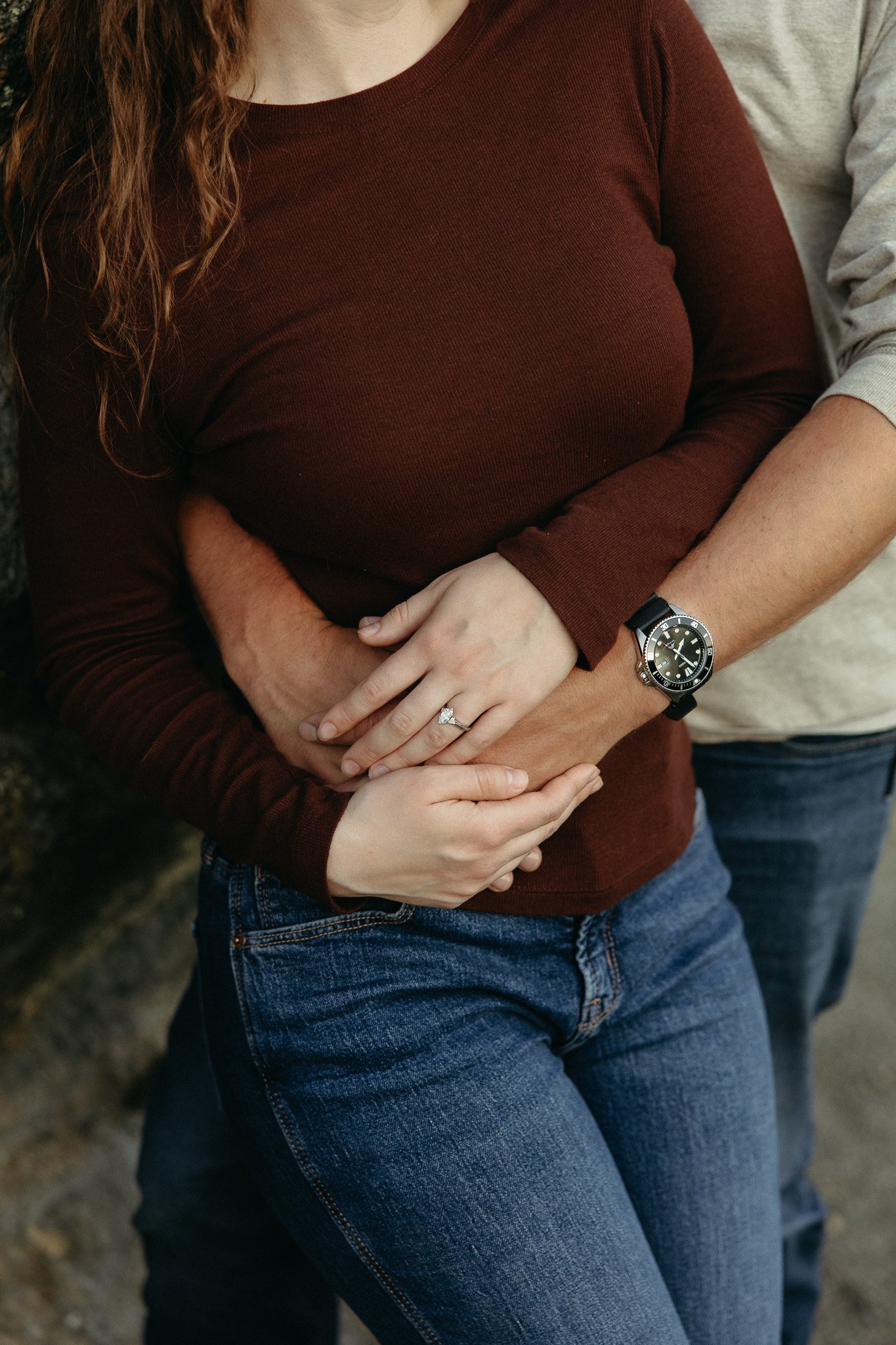 A woman with long wavy red hair, wearing a burgundy long-sleeve shirt and blue jeans, is embracing a man from behind, with both of their hands resting on her stomach. The man is wearing a gray long-sleeve shirt and a wristwatch. The woman's ring is v