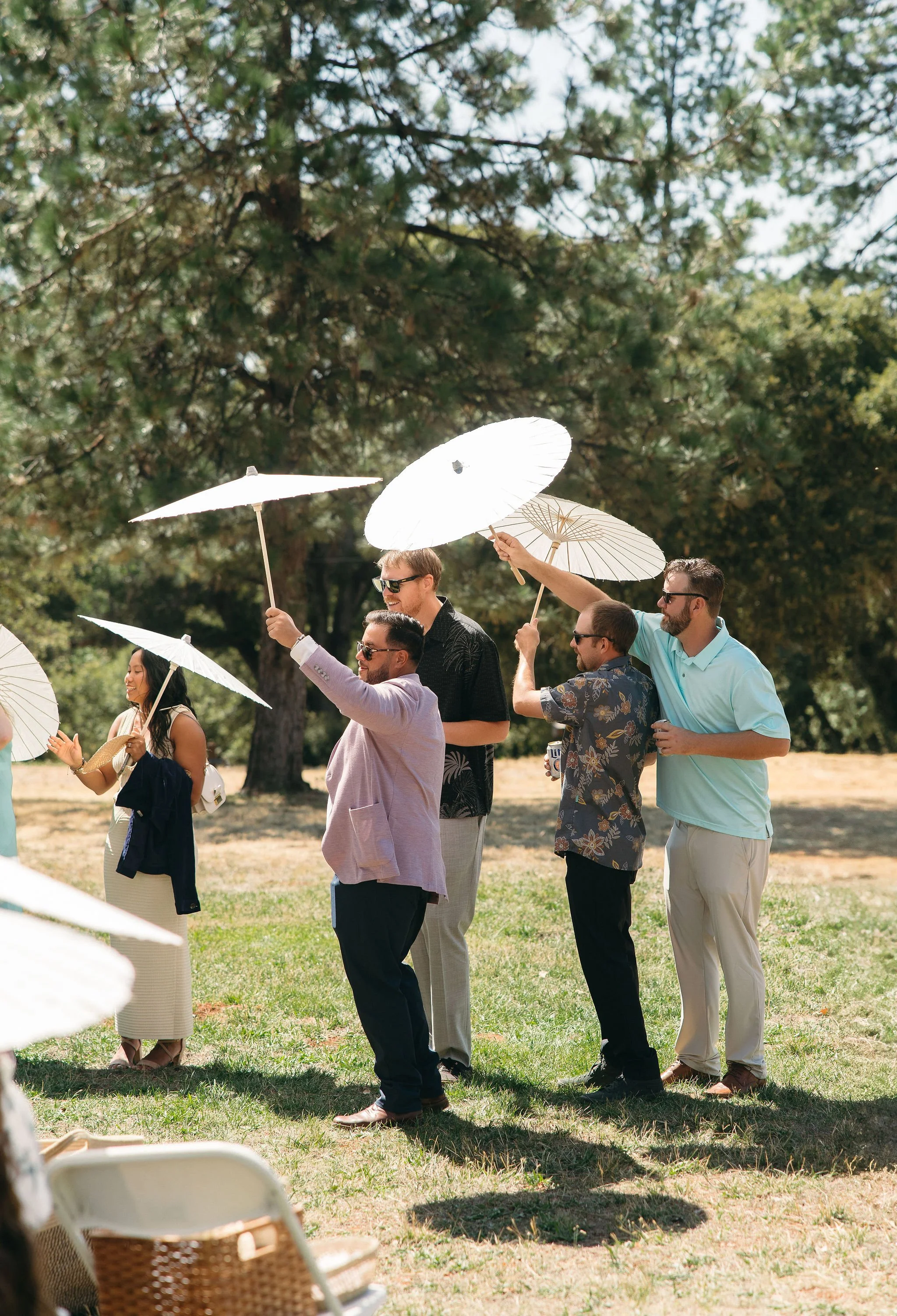 A group of people at an outdoor gathering holding white parasols, enjoying sunny weather in a park with trees.