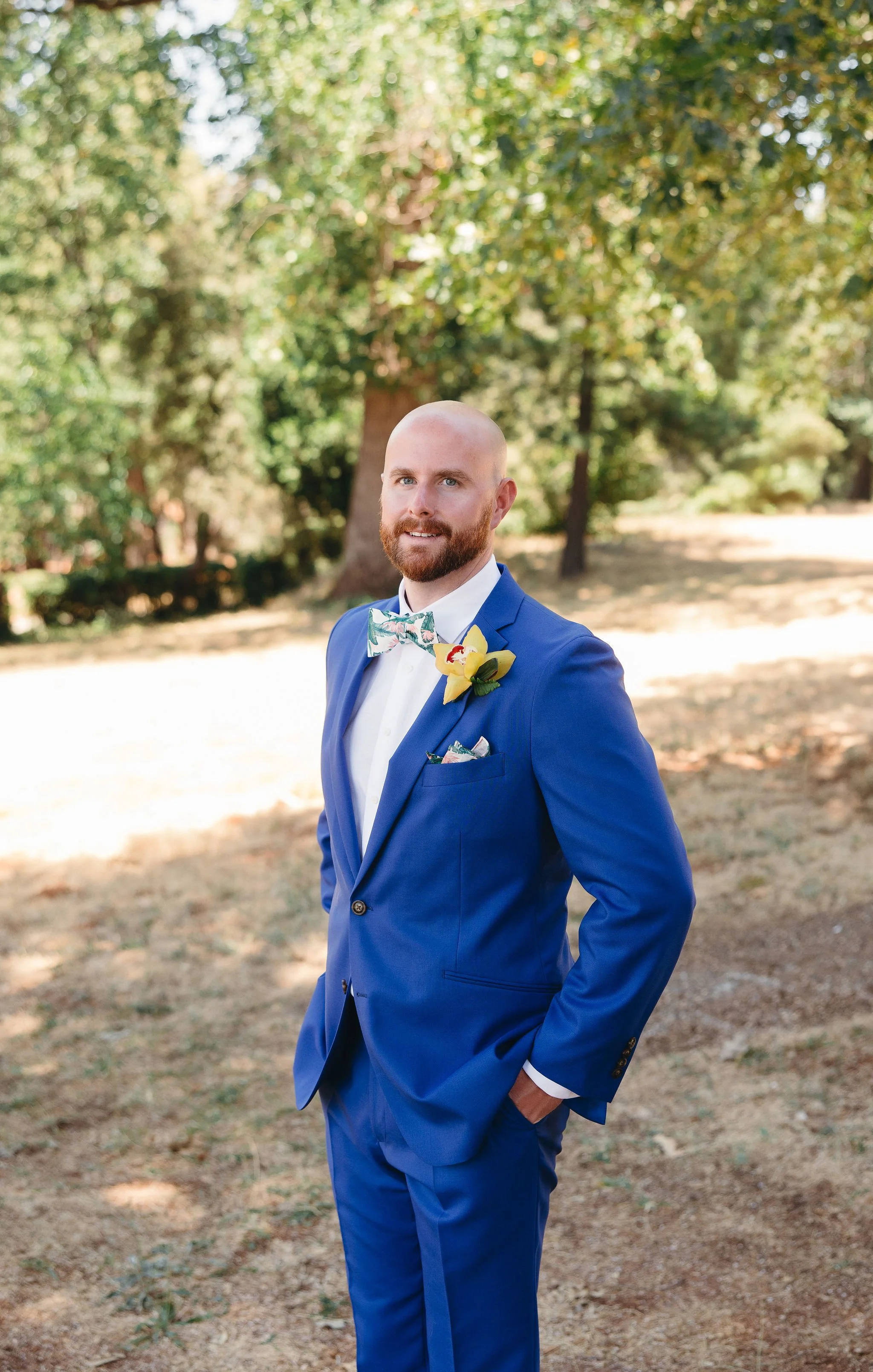 A man dressed in a blue suit with a floral bow tie and a yellow flower boutonniere, standing outdoors in a wooded area with sunlight filtering through the trees.