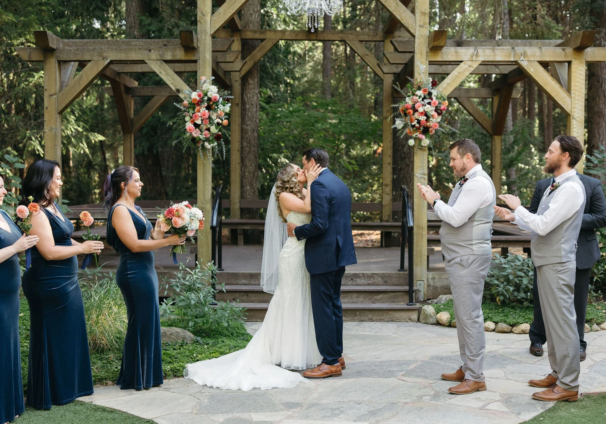 A wedding ceremony outdoors with the bride and groom kissing, surrounded by bridesmaids and groomsmen in a wooded area with floral decorations.