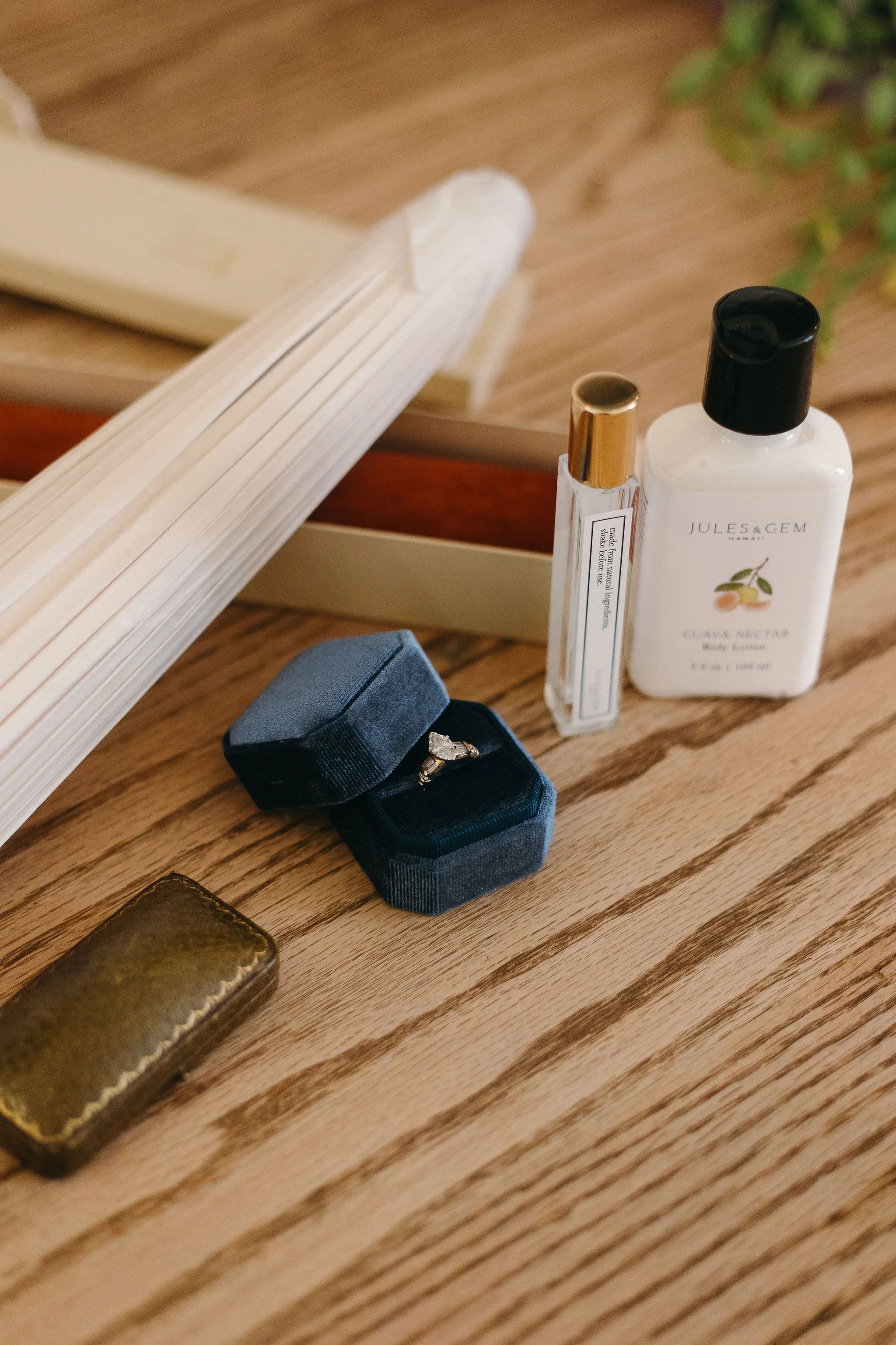 A wooden tabletop with a small jewelry box holding an engagement ring, a gold-colored wallet, bottles of body lotion and perfume, and a mirror.