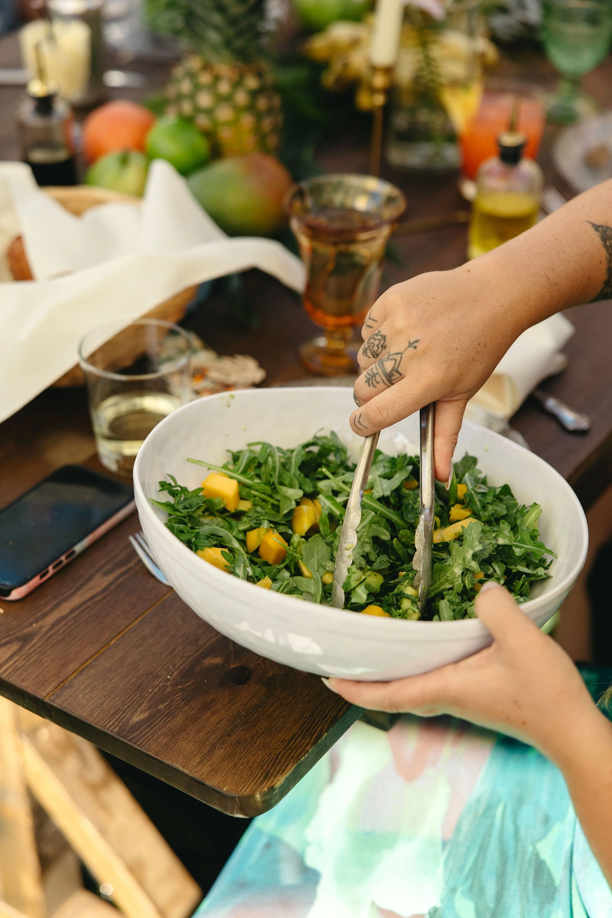 Person tossing a fresh green salad with mango chunks in a white bowl at a table with various drinks, fruits, and a mobile phone.