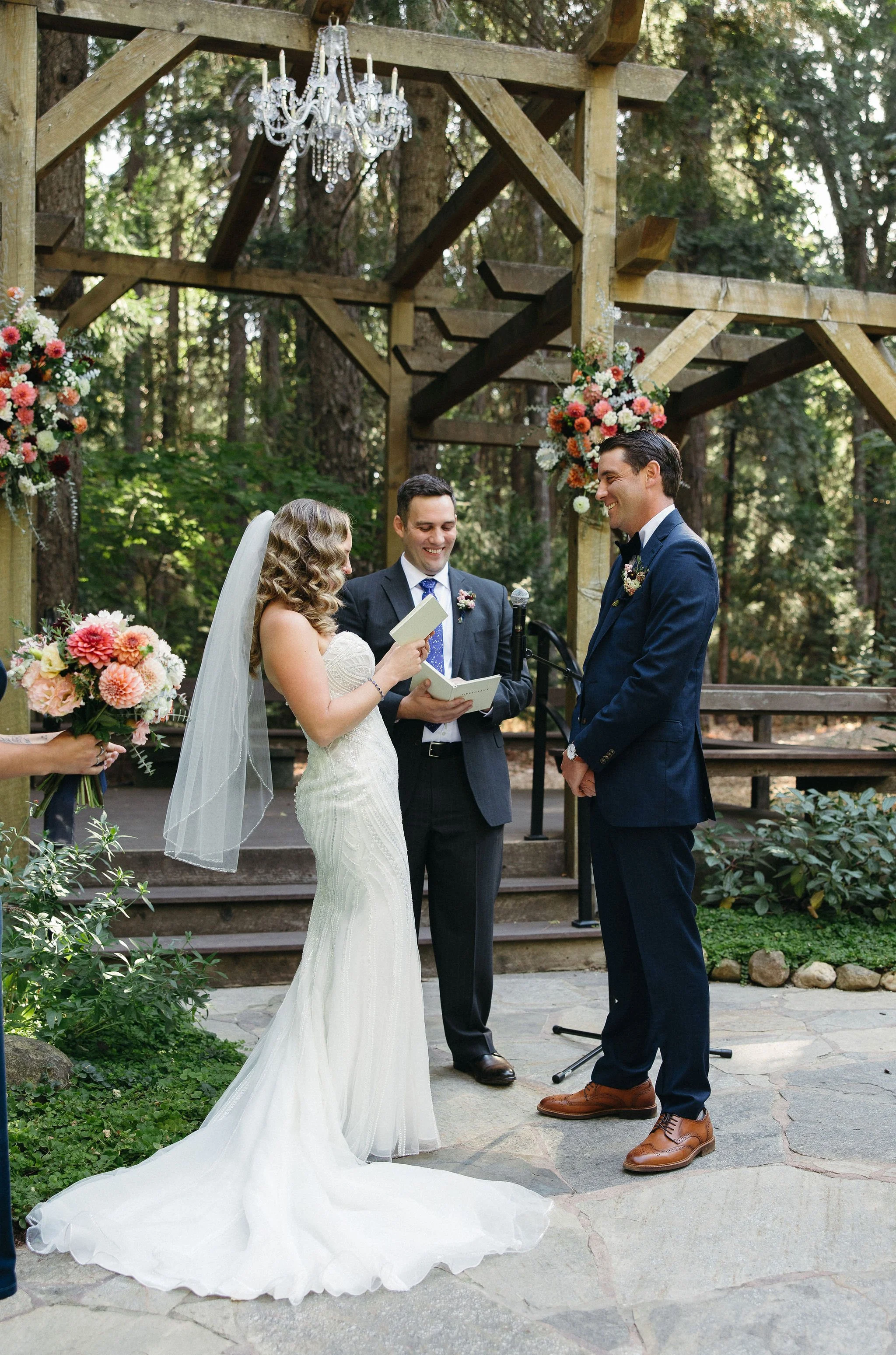 A wedding ceremony taking place outdoors in a forested area with a wooden arch decorated with pink and white flowers, a chandelier hanging above, and a bride and groom exchanging vows with an officiant present.