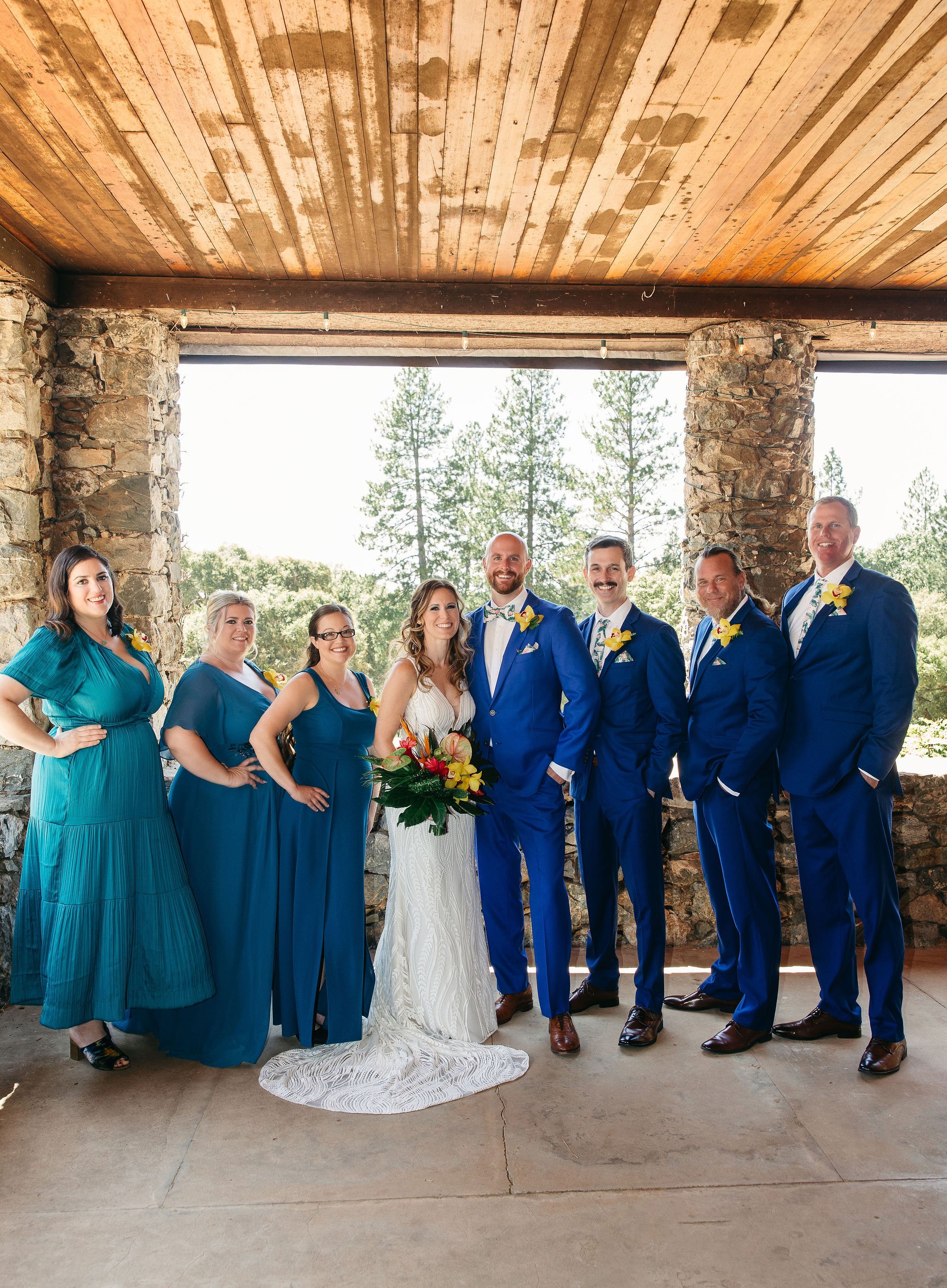 A wedding party stands together outdoors under a stone and wooden pavilion, with trees and blue sky in the background. The group includes a bride in a white gown holding a colorful bouquet, seven men in blue suits, and three women in blue dresses.