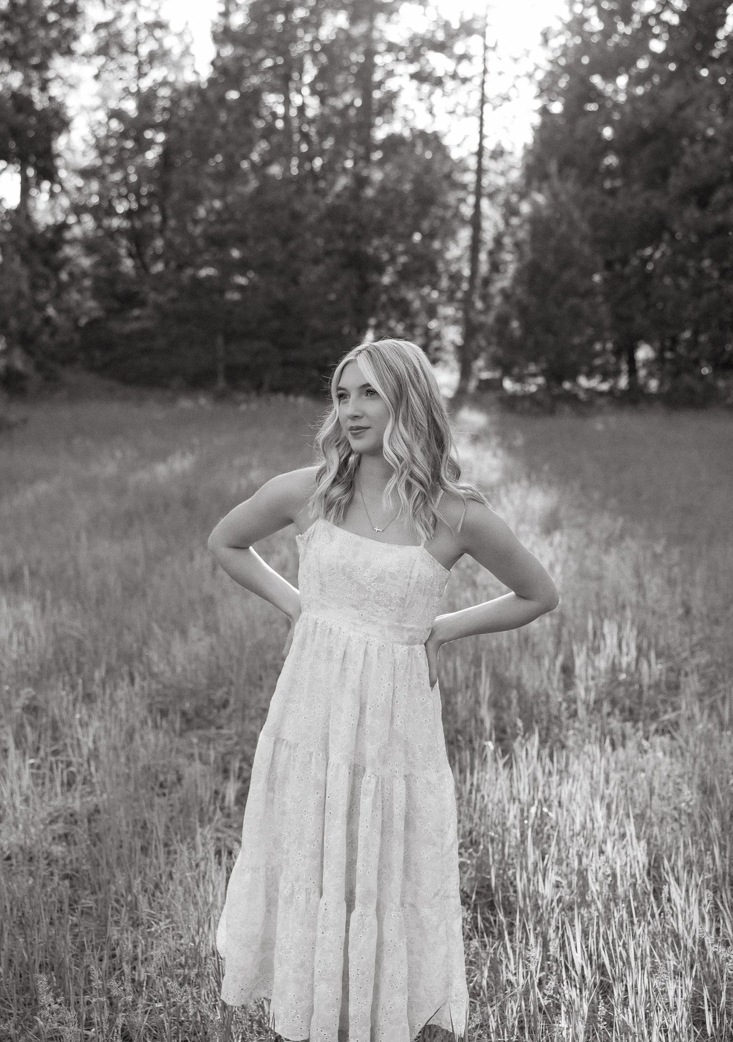 A woman in a white lace dress standing in a grassy field with trees in the background, looking to the side with her hands on her hips.