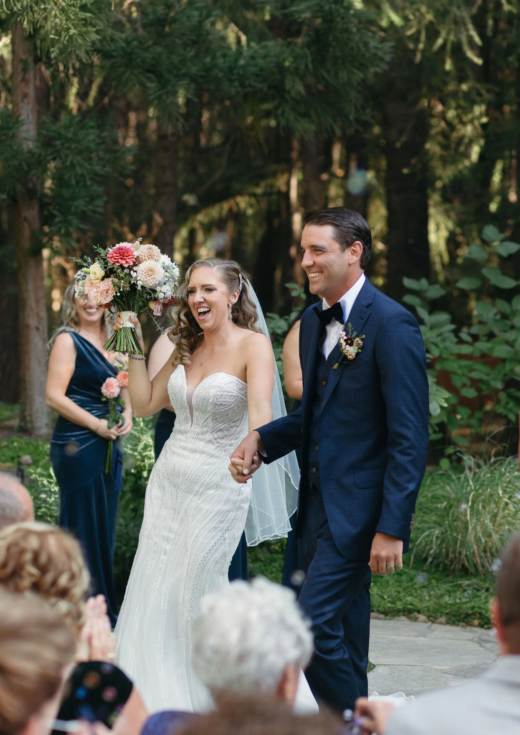 Happy bride and groom holding hands during their outdoor wedding ceremony in a forest setting, surrounded by guests and bridesmaids, with the bride holding a bouquet of pink and white flowers.