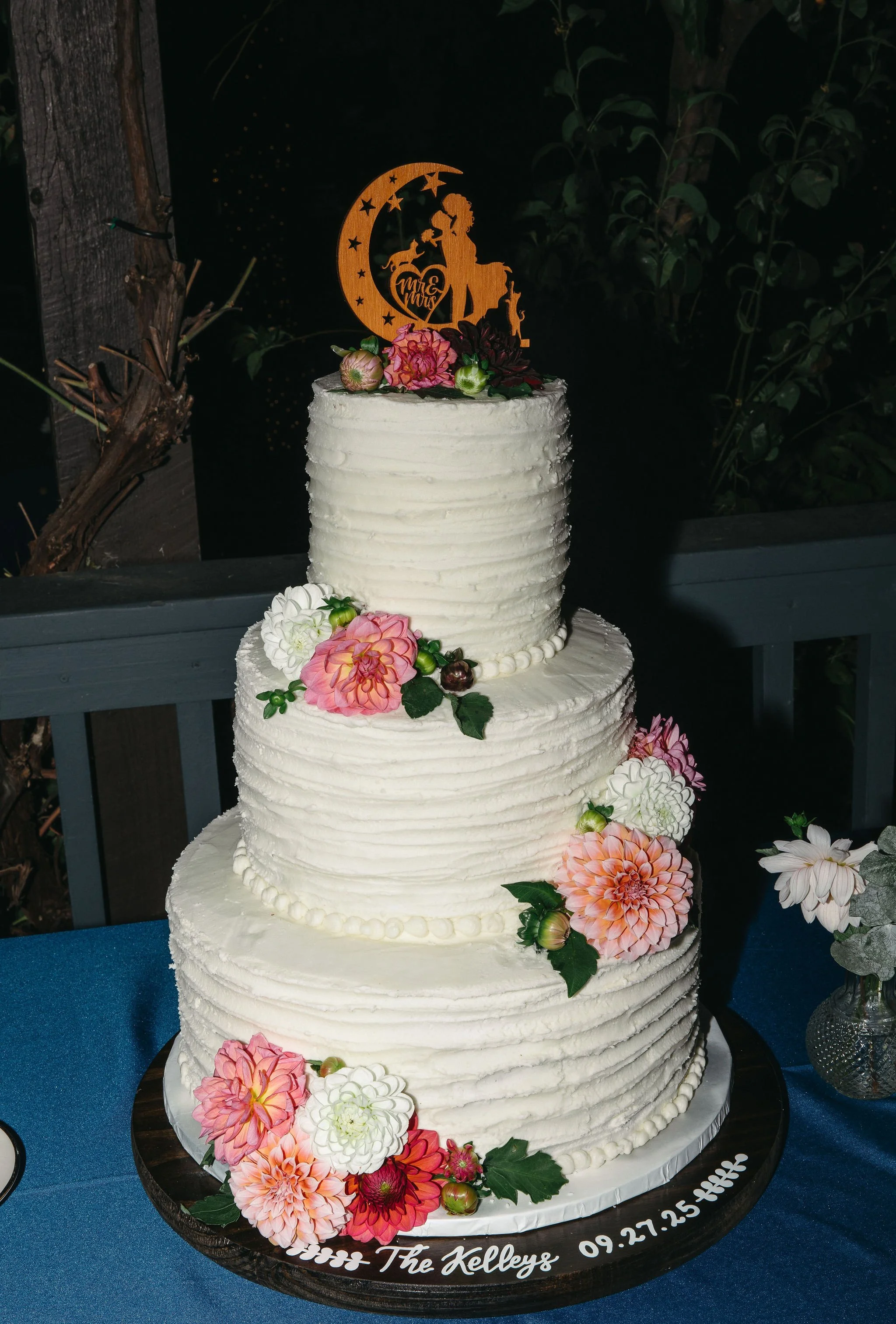 Three-tiered wedding cake decorated with colorful flowers and a wooden topper that reads 'Mr. & Mrs.', with the inscription 'The Kellys' and the date '09.27.25' on the base.