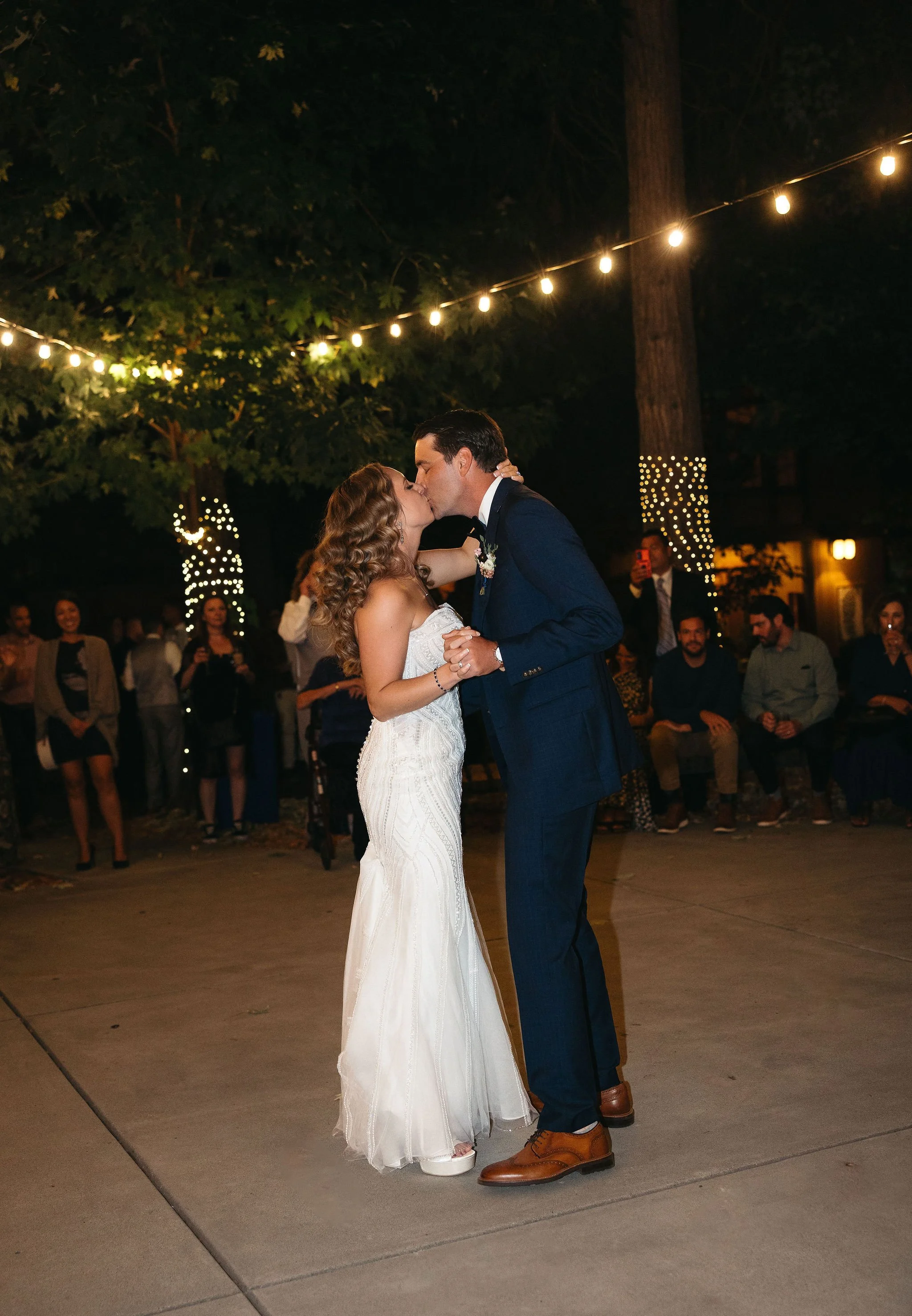 A bride and groom share a kiss during their wedding reception under string lights and tall trees with string lights wrapped around the trunks. Guests watch in the background.