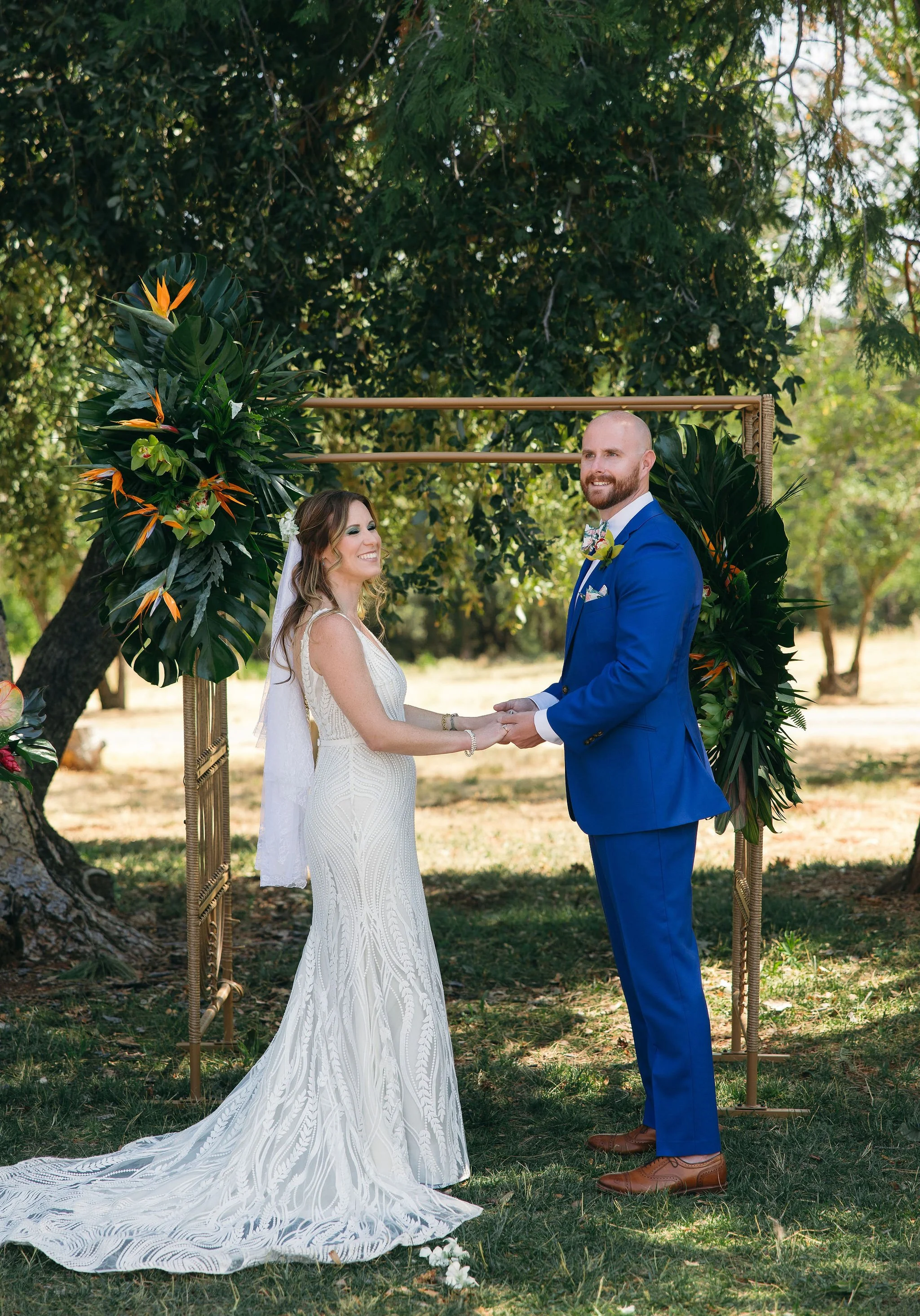 A bride and groom holding hands and smiling at each other during an outdoor wedding ceremony, standing under a decorative arch with tropical greenery and flowers, in a lush, sunlit garden.