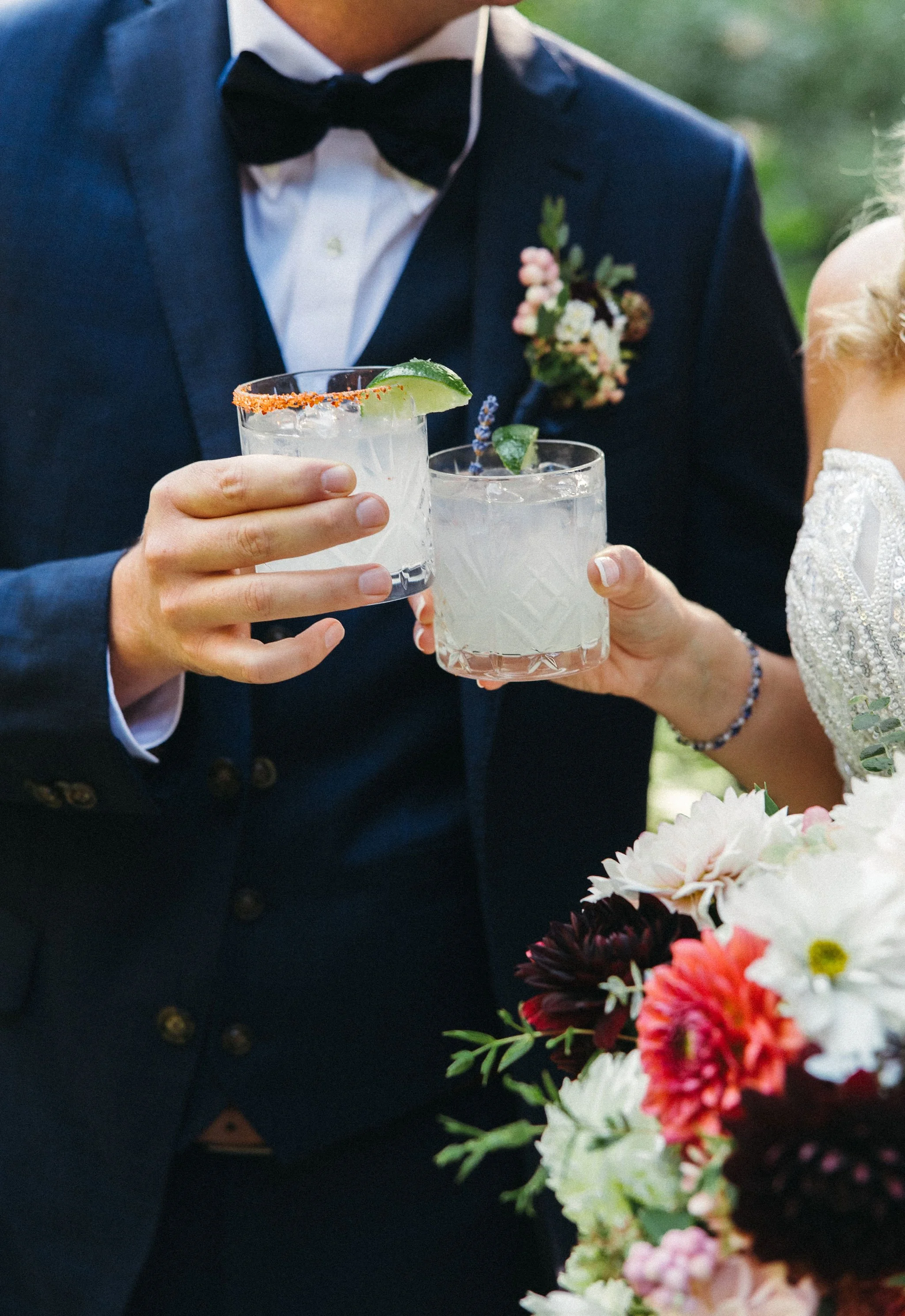 Man and woman in wedding attire toasting with cocktails outdoors.
