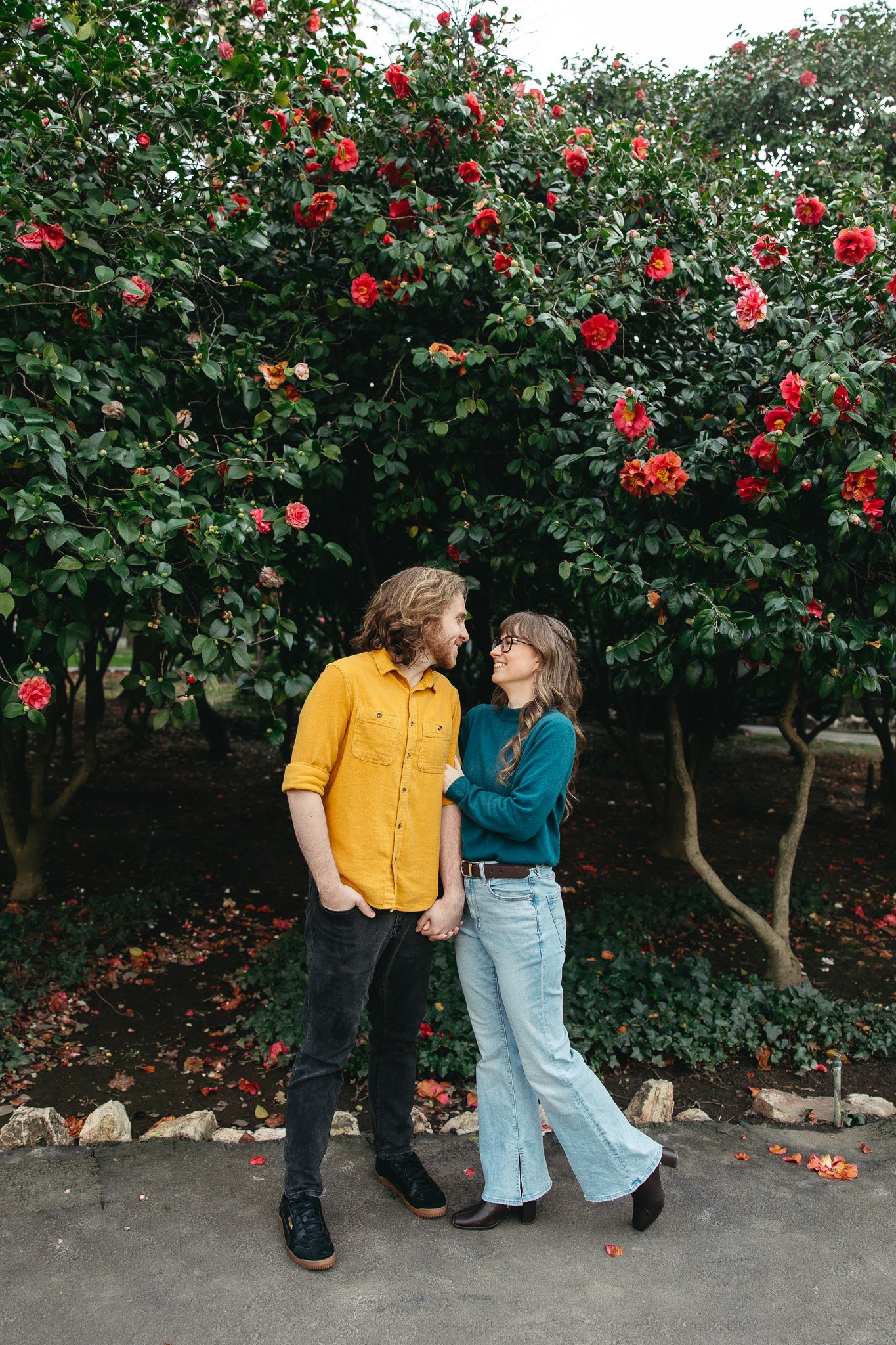 A couple holding hands and smiling at each other under a flowering bush with pink and red flowers.