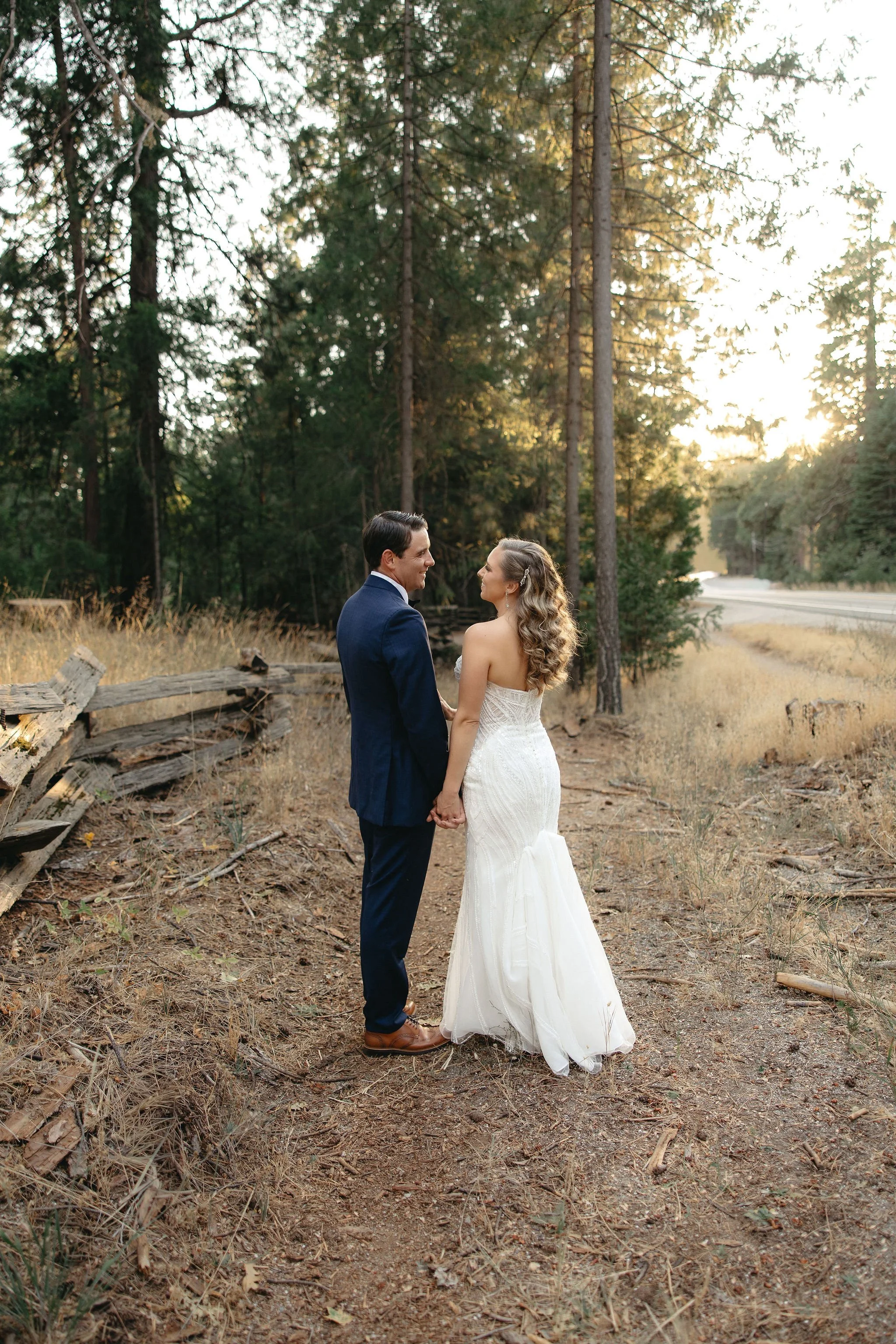A newlywed couple holding hands and gazing at each other on a forest trail at sunset, with trees and a dirt path in the background.