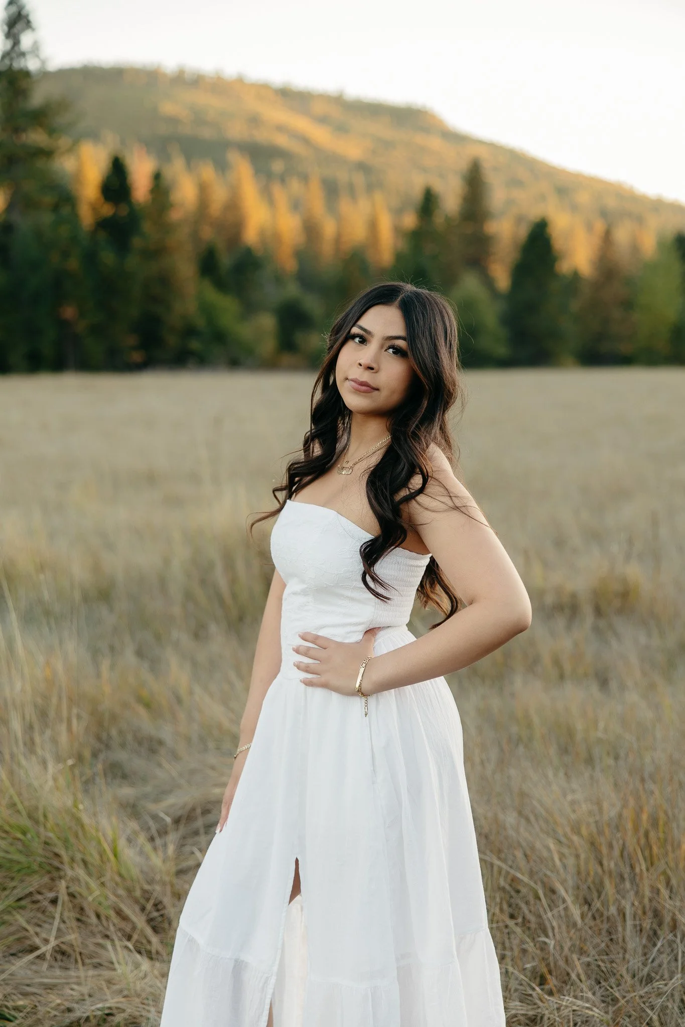 A young woman with long wavy dark hair wearing a white strapless dress stands in a field of tall grass with a backdrop of trees and a mountain, during golden hour.