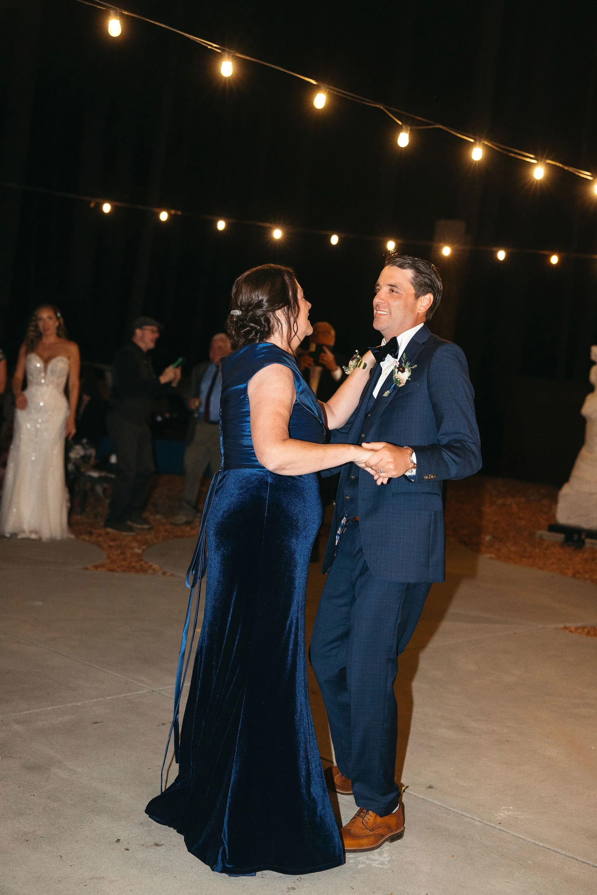 A couple dancing at a wedding reception outdoors at night, illuminated by string lights, with other guests watching in the background.