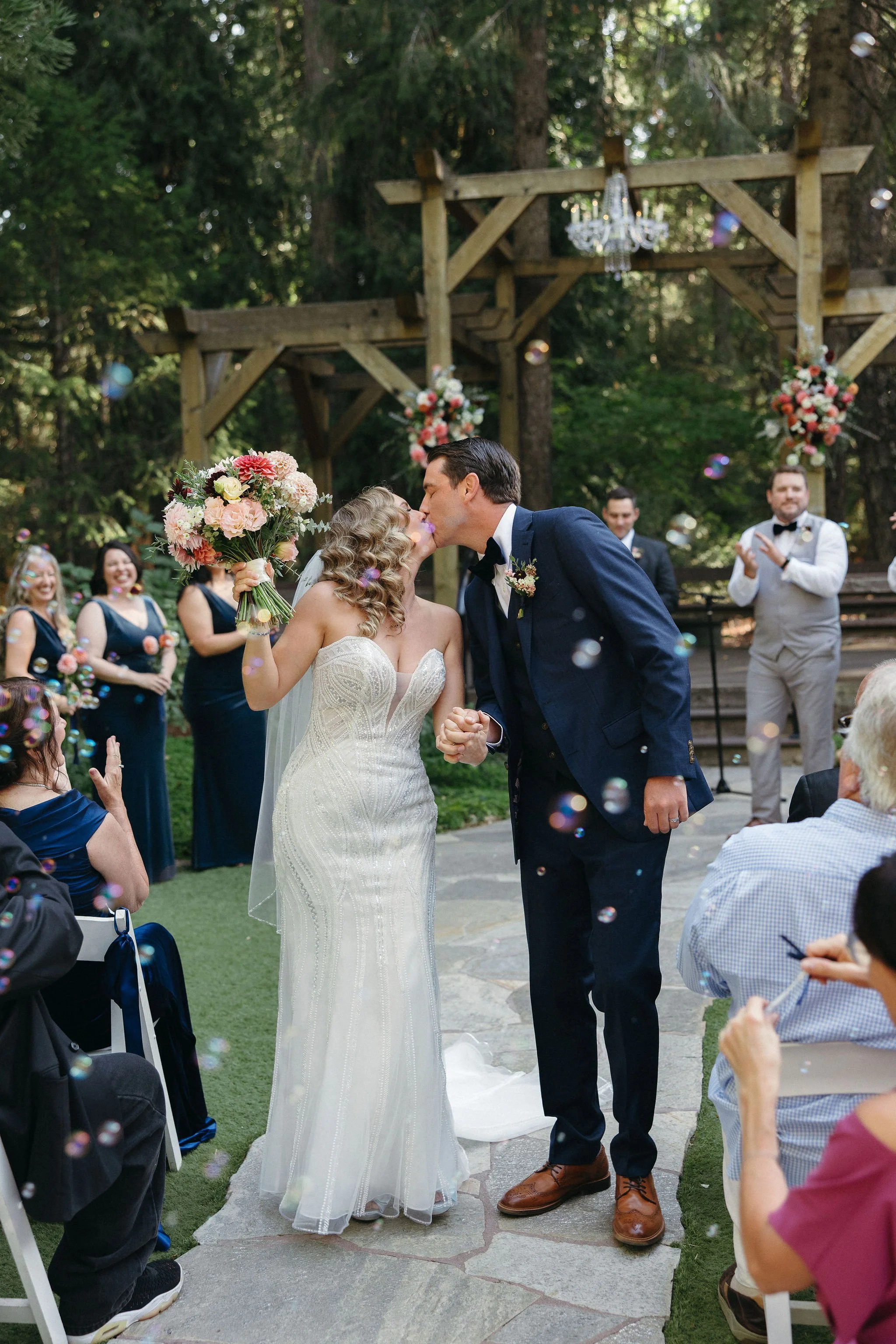Bride and groom sharing a kiss during their outdoor wedding ceremony, surrounded by friends and family, with floral decorations and a wooden arch in a wooded area.