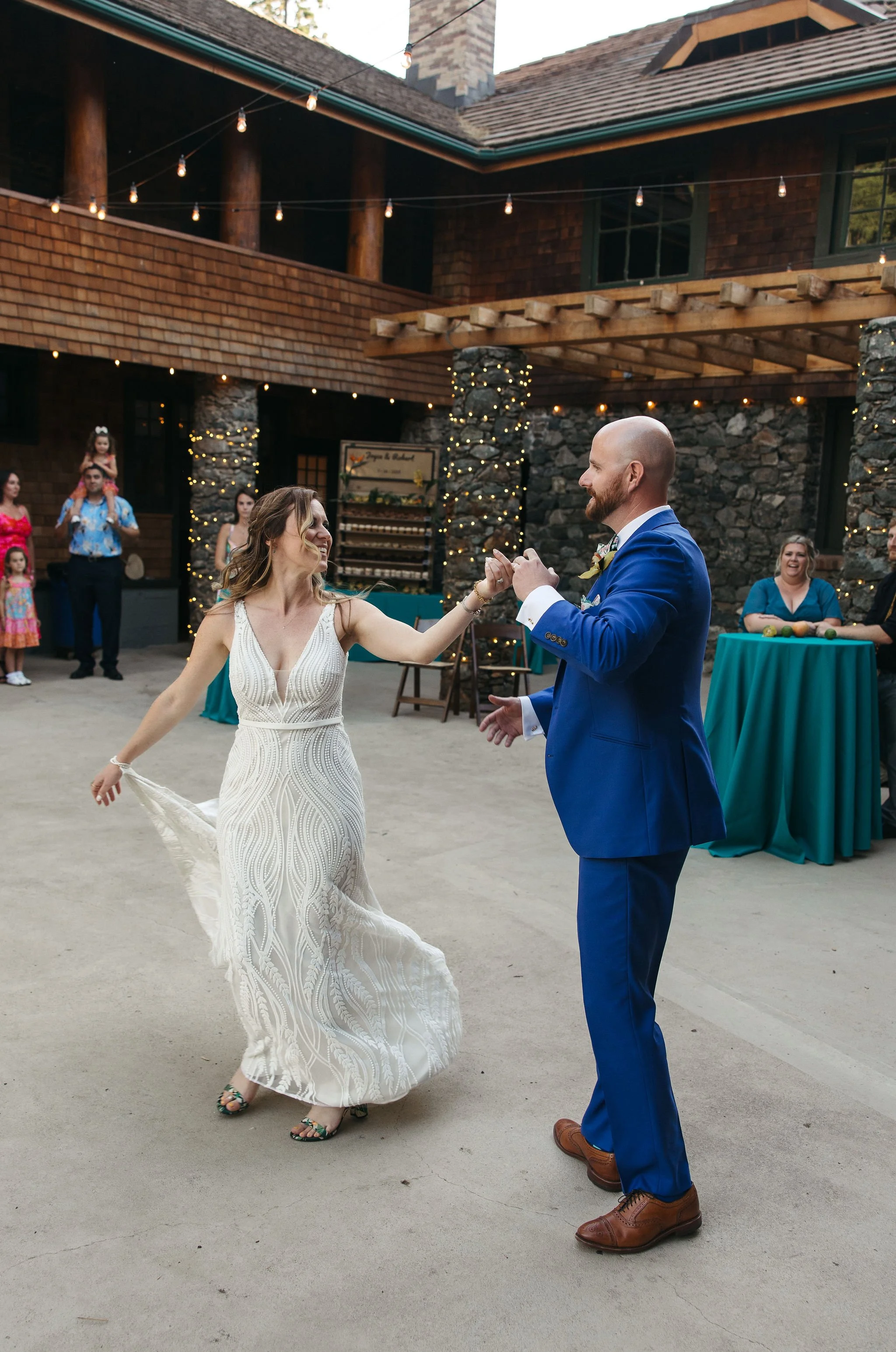 A bride and groom dancing at their wedding reception outdoors, with guests and string lights in the background.