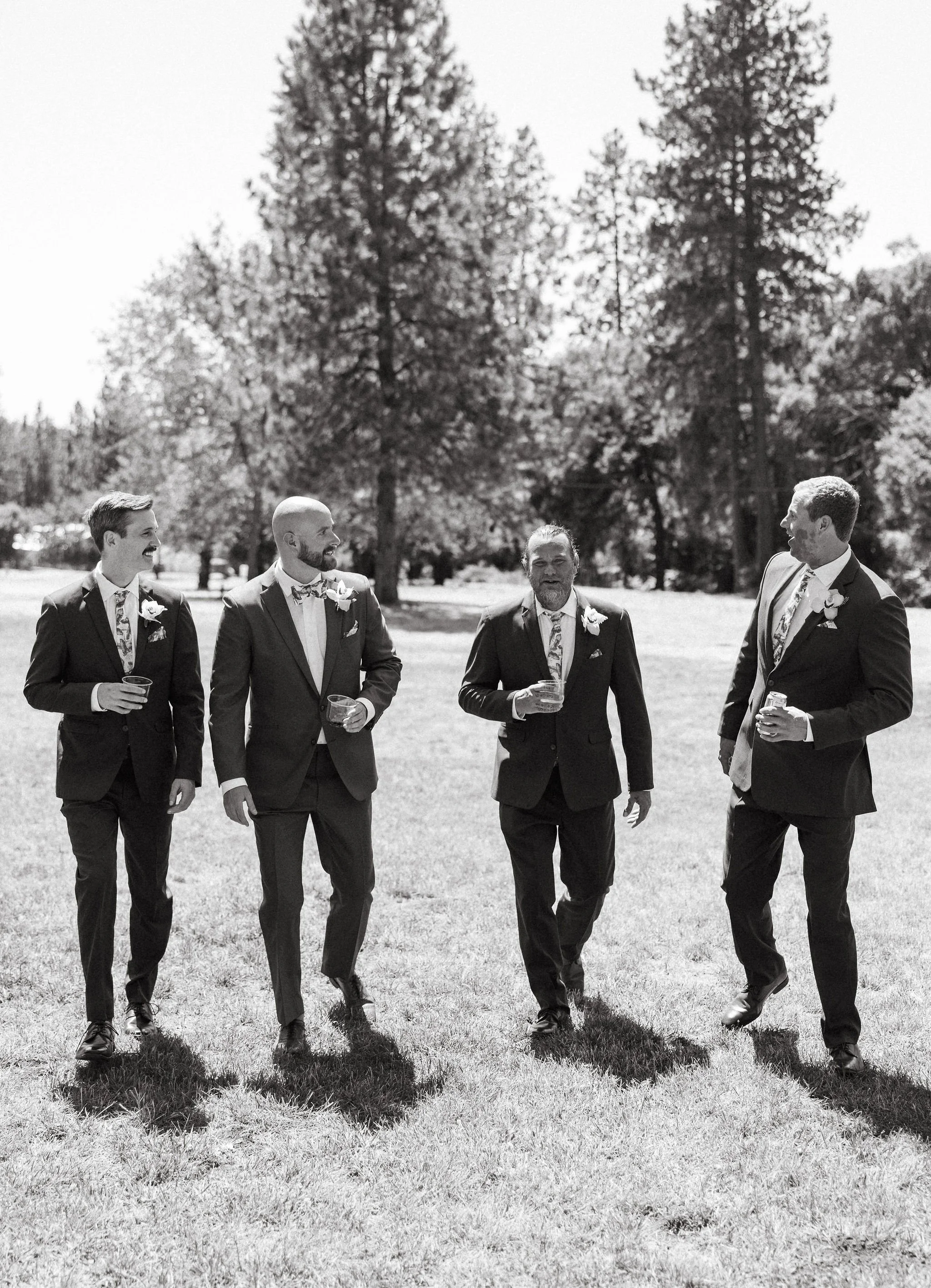 Four men in suits walking on grass in a park, holding drinks, with trees in the background.