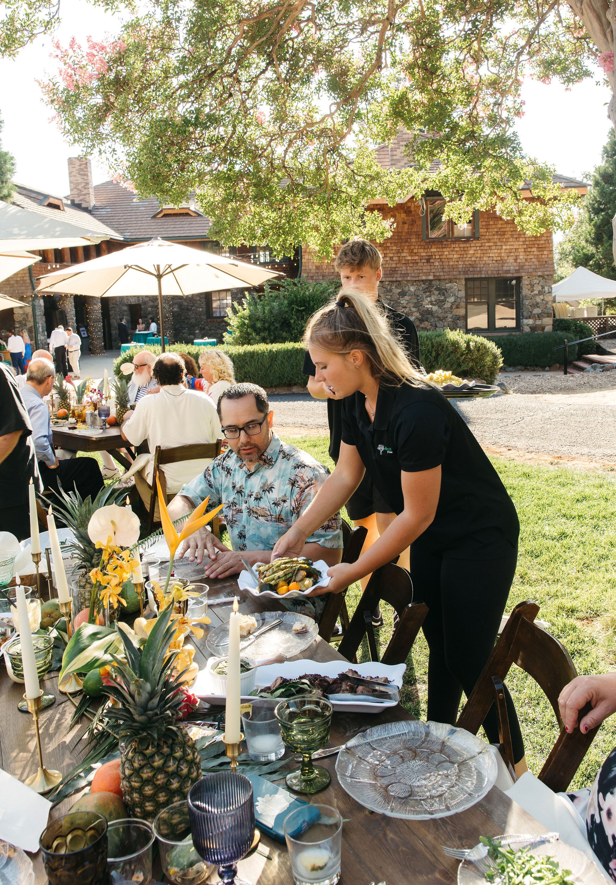 People enjoying an outdoor gathering with food and decorations under a large tree on a sunny day.