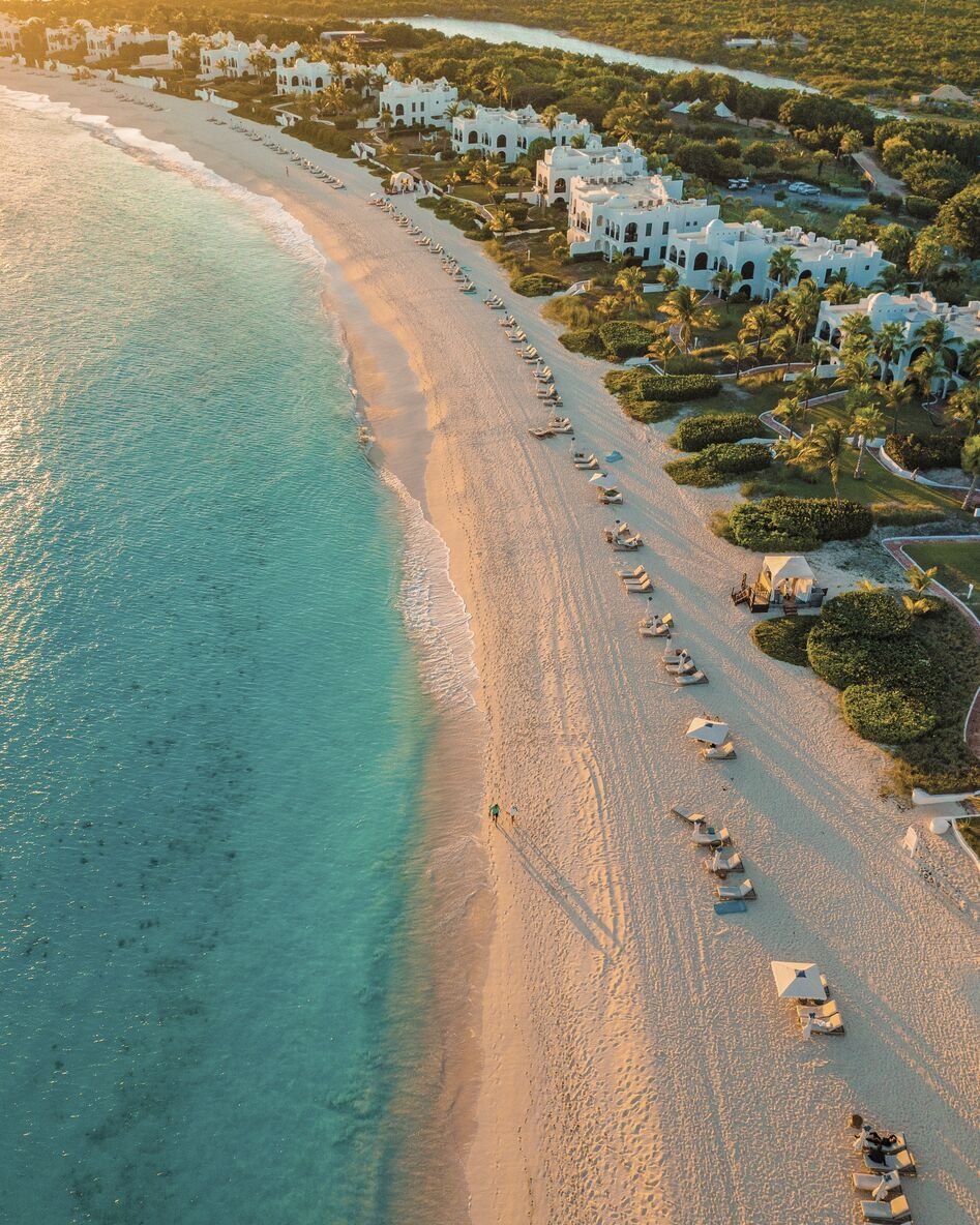Aerial view of a tropical beach lined with white resort buildings, palm trees, lounge chairs, and umbrellas, with calm turquoise water and a sandy shoreline.