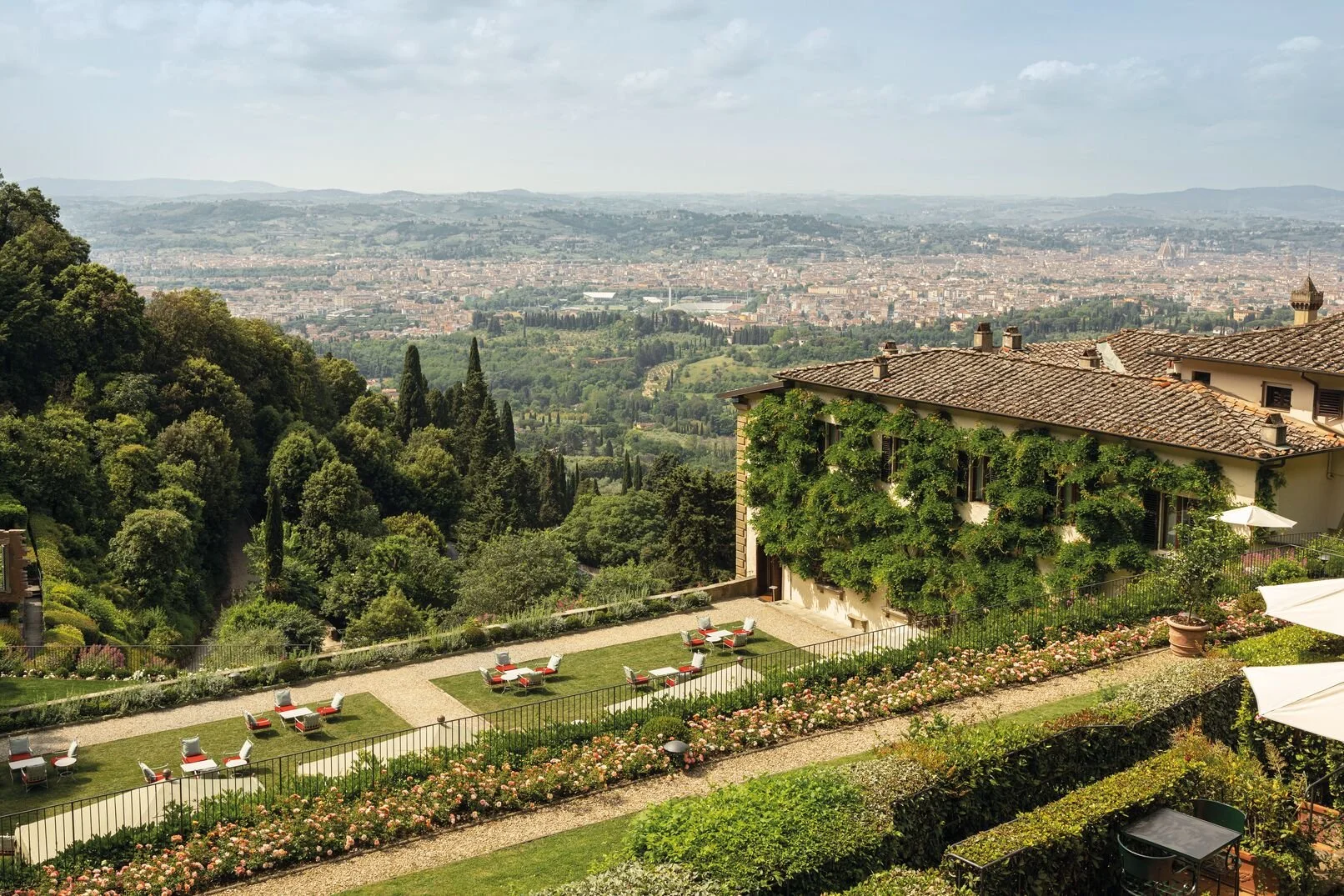 Scenic view of a lush green hillside garden with patio furniture and a building covered in greenery, overlooking the cityscape of Florence and rolling hills in the distance under a partly cloudy sky.