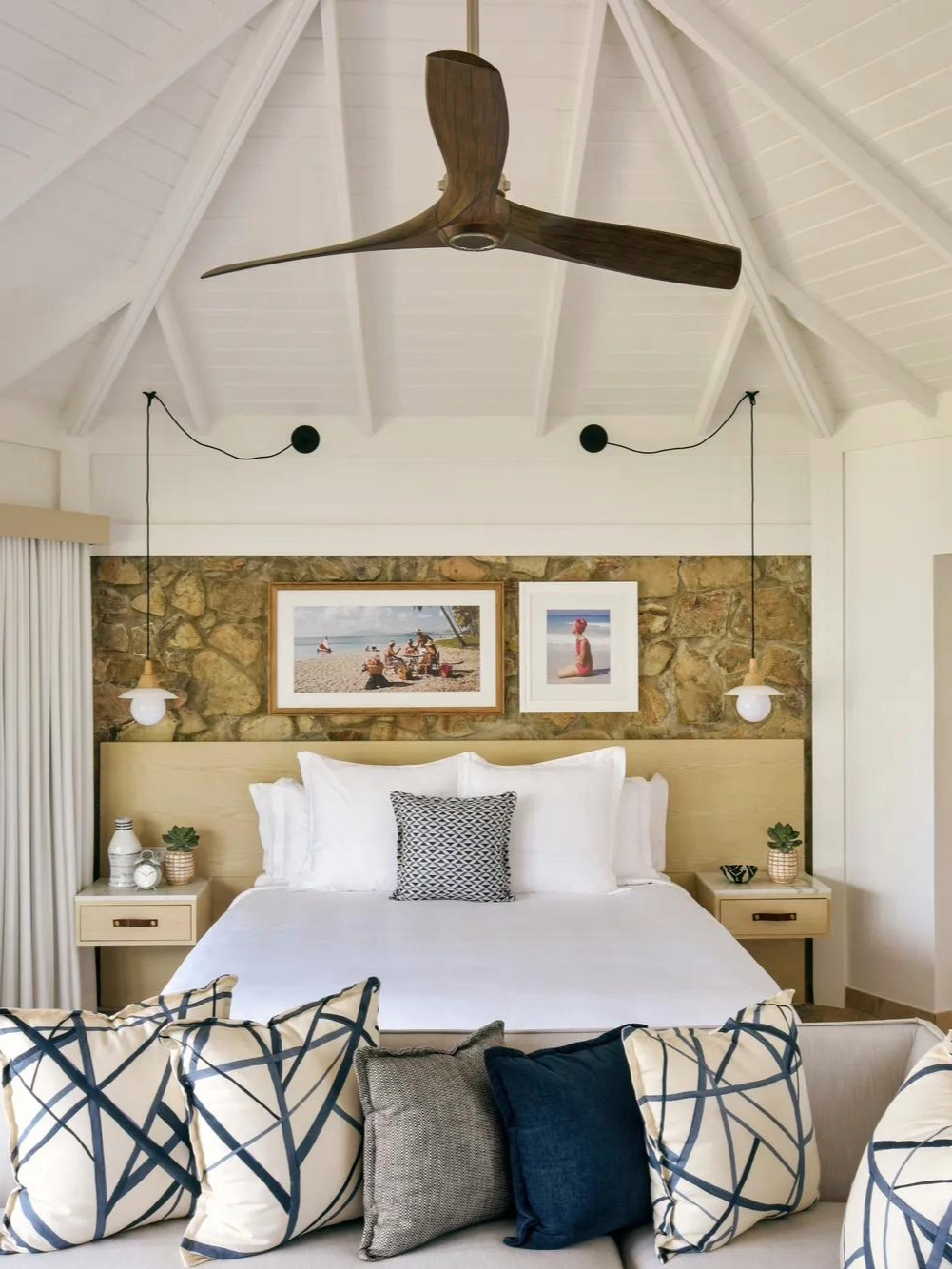 A bedroom with a white bed, decorative pillows, a stone accent wall, and two framed beach photos above the bed. There are wall-mounted pendant lights, a ceiling fan, and side tables with plants.