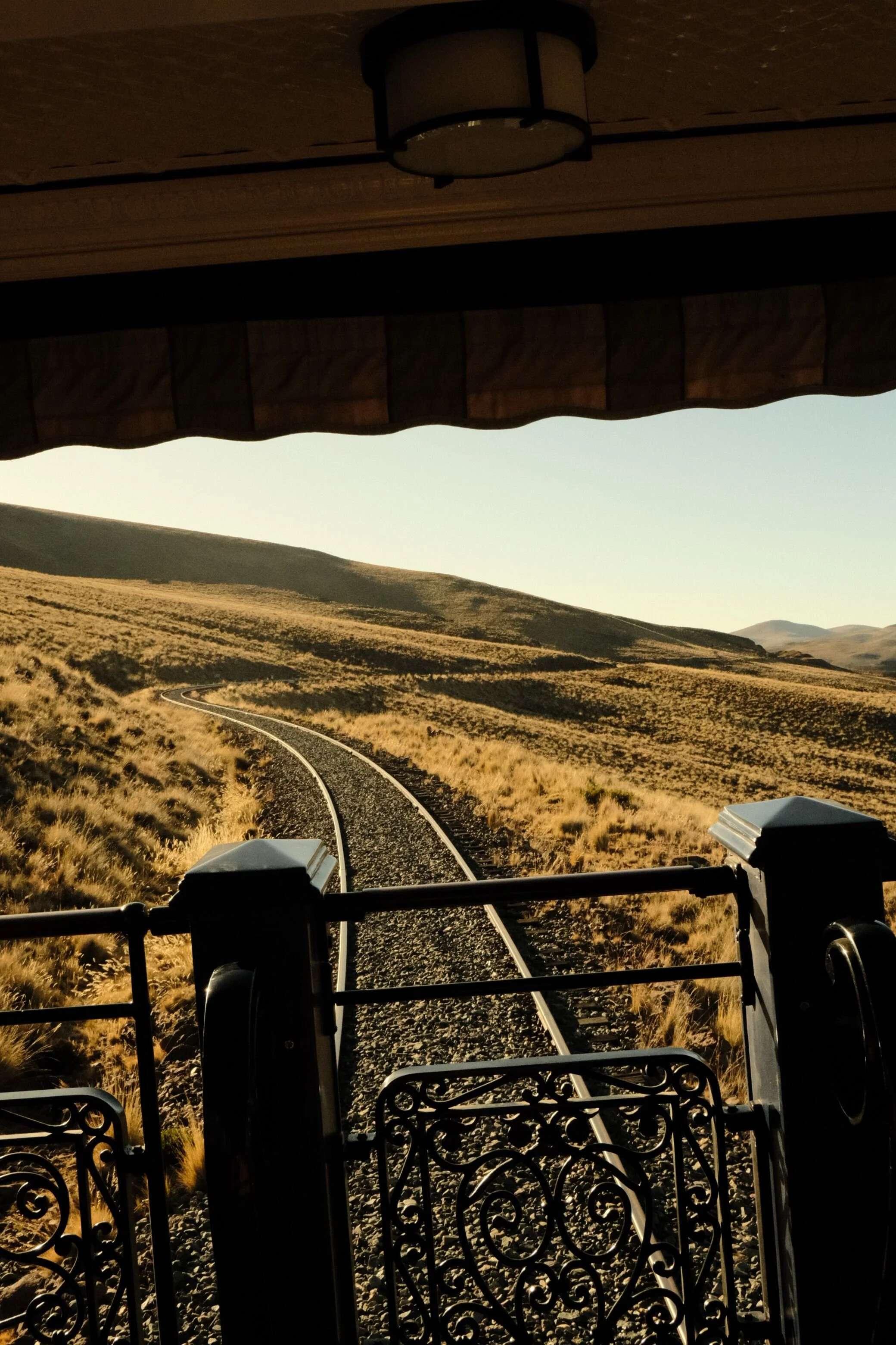 View from a train or tram showing a curved railway track through a dry, hilly landscape with grasses and shrubs, under a partially covered roof structure with a lamp fixture overhead.