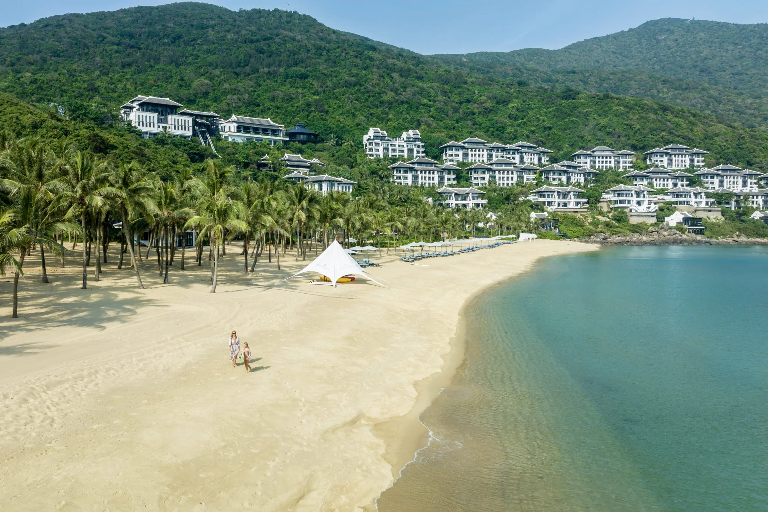 Beach with people walking on it and villas dotted amongst palm trees