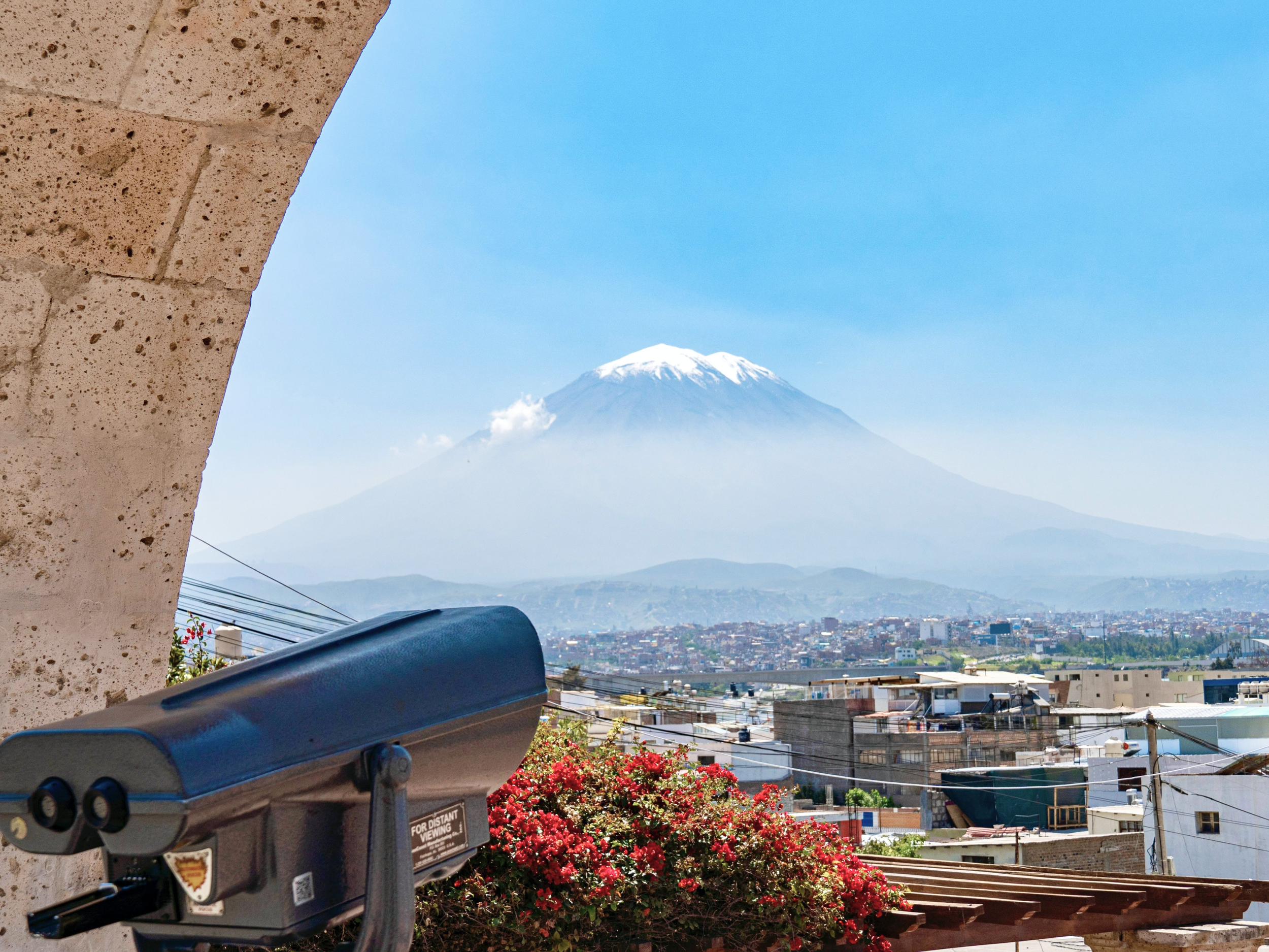 Volcano El Misti viewed from a scenic lookout in Arequipa, Peru, on a clear blue-sky day, with blooming bougainvillea and a viewing telescope in the foreground.