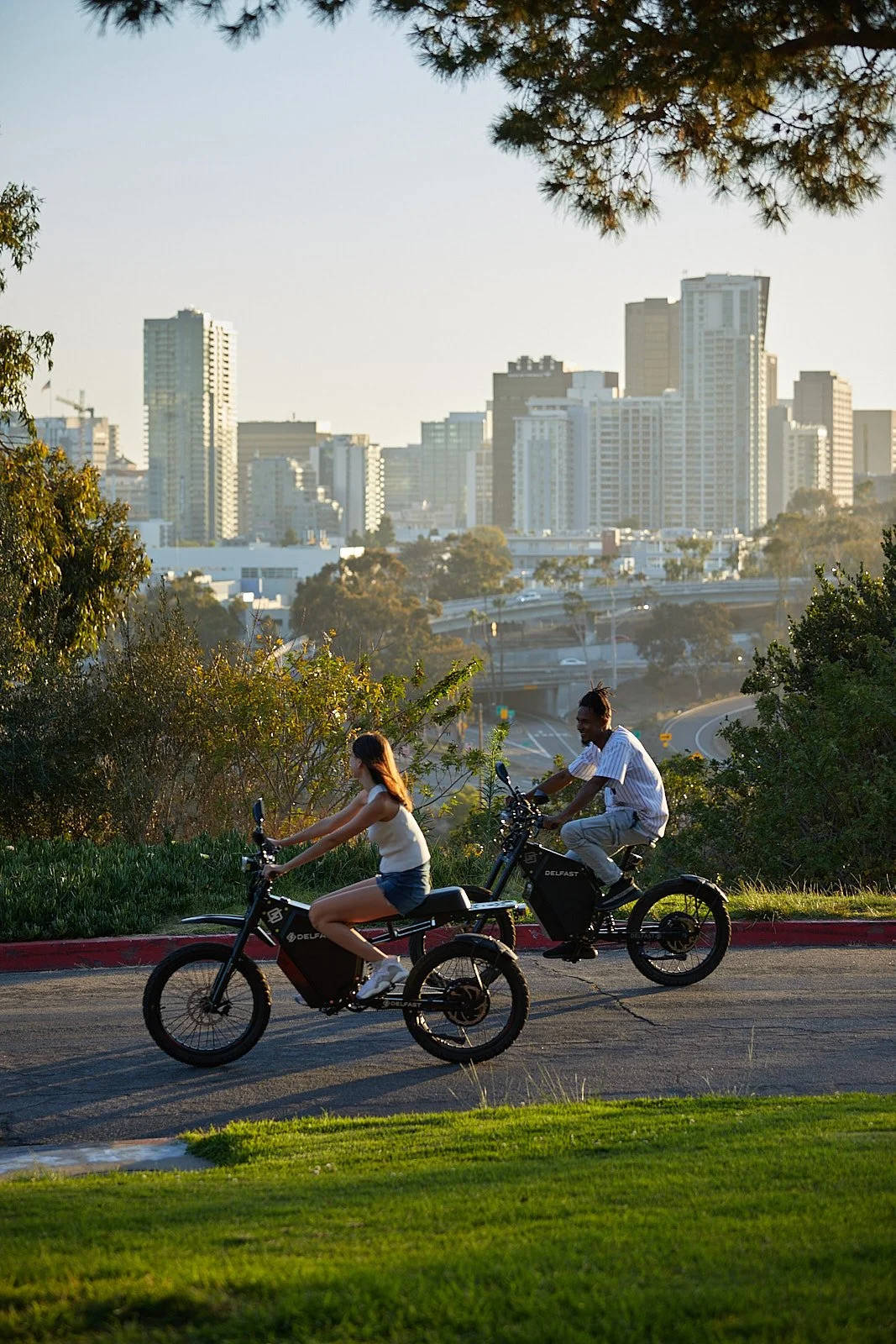 A man and a woman riding black electric bikes along a paved road at sunset, with a dense city skyline and skyscrapers in the background.