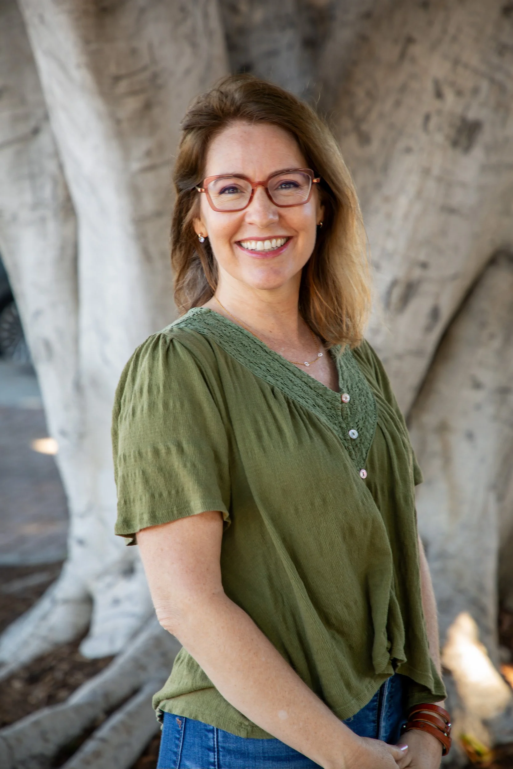 A woman with shoulder-length brown hair, glasses, and earrings smiling outdoors wearing a green blouse and blue jeans, with a large tree trunk and branch in the background.