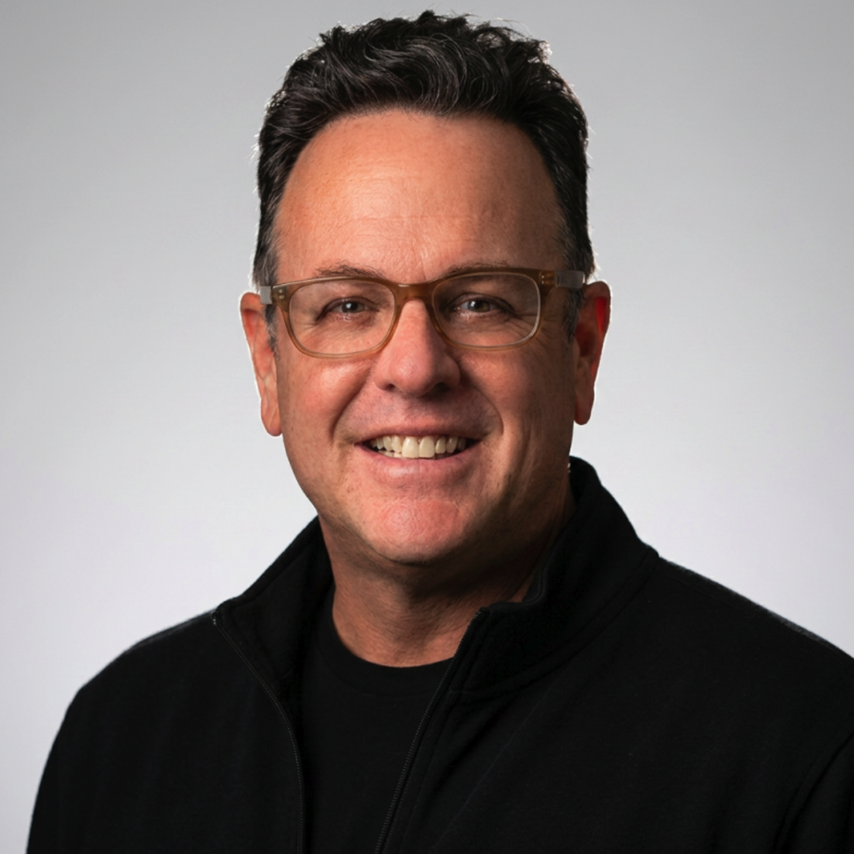 A smiling man with brown hair and glasses wearing a black collared shirt on a neutral plain light gray background.