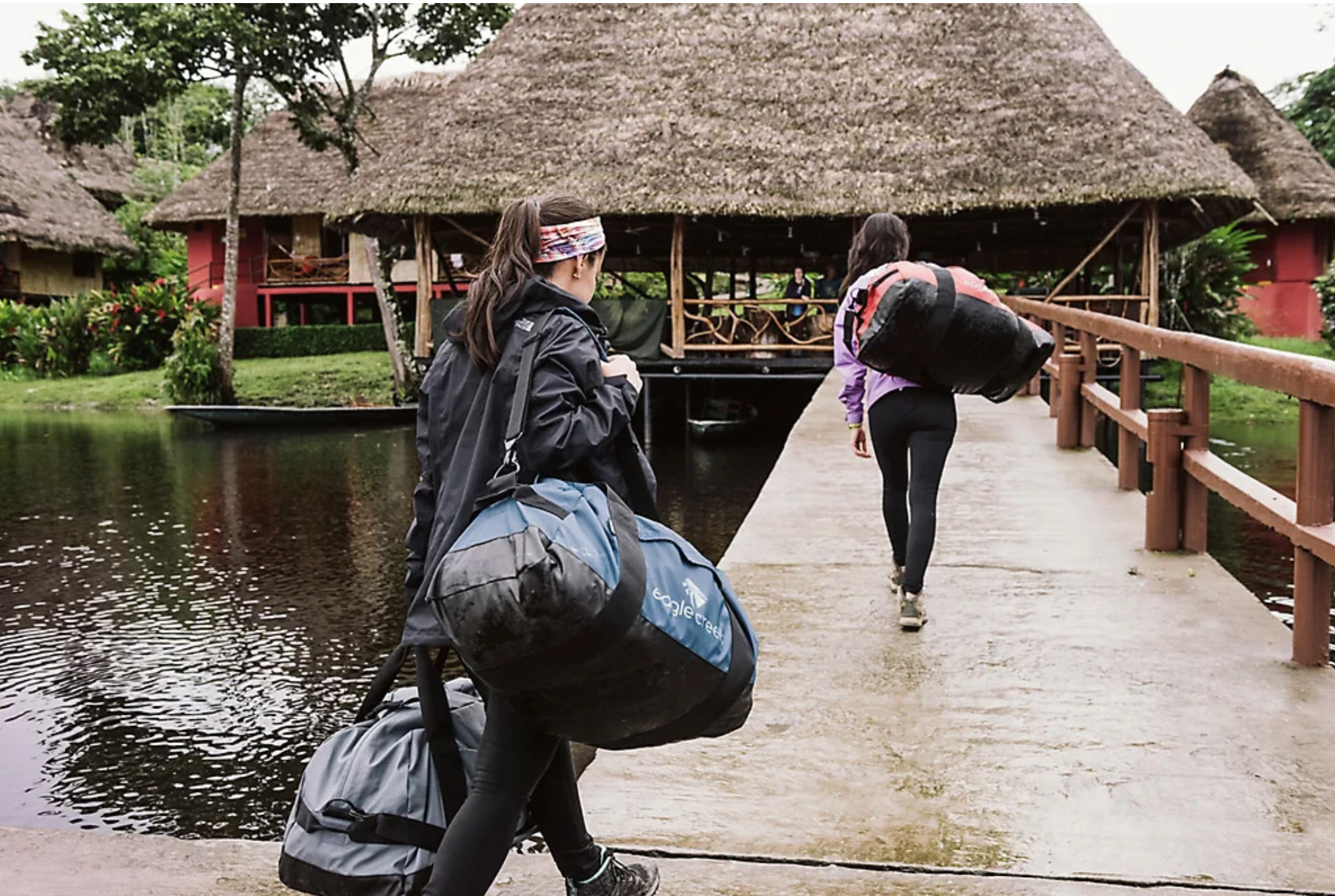 Two travelers carrying large Eagle Creek duffel bags across a wet dock toward a tropical thatched-roof lodge by the water.