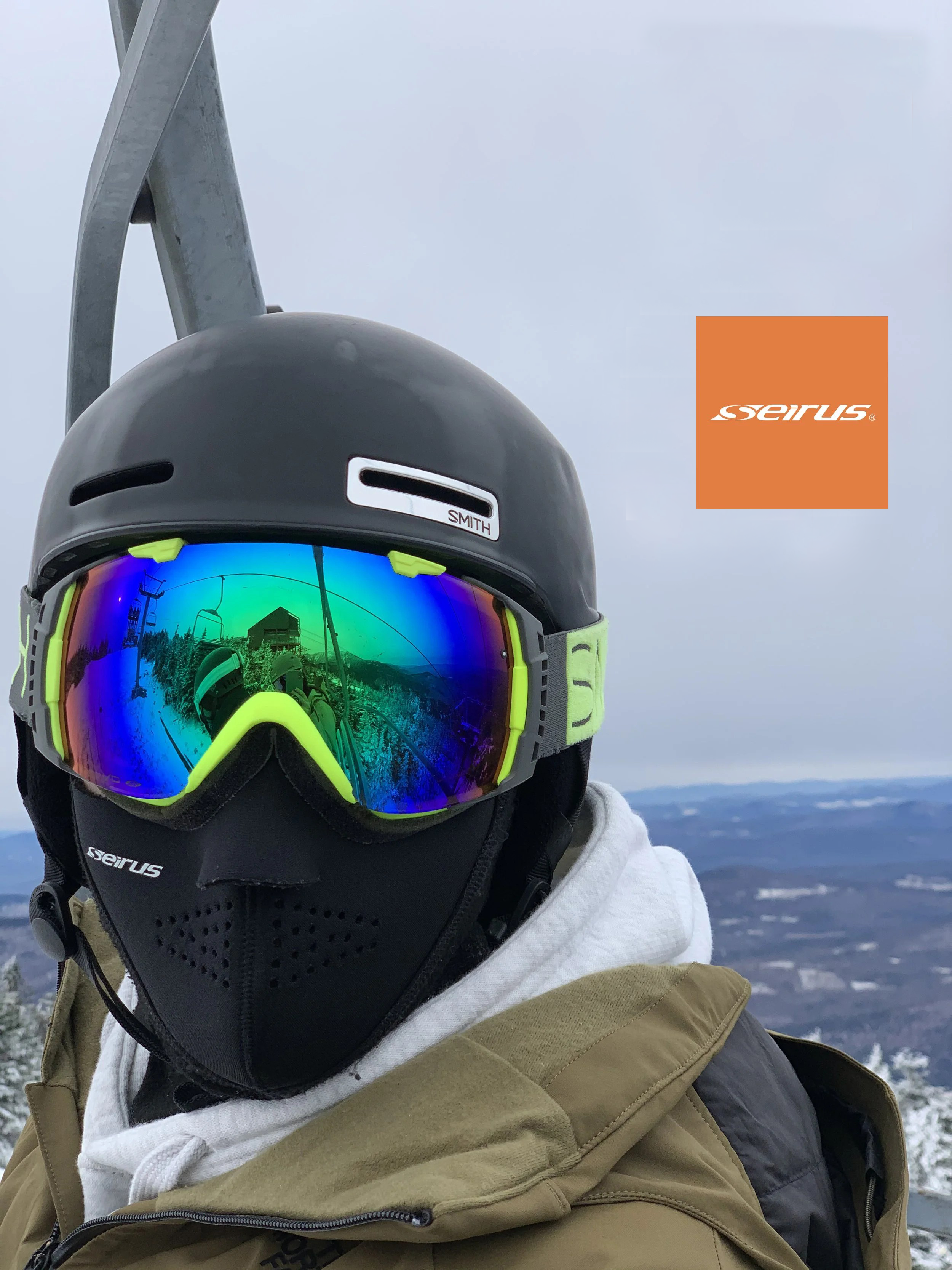 Close-up of a skier wearing a black Seirus Neofleece face mask, a black Smith helmet, and neon green Smith goggles reflecting a ski lift and mountain scenery.
