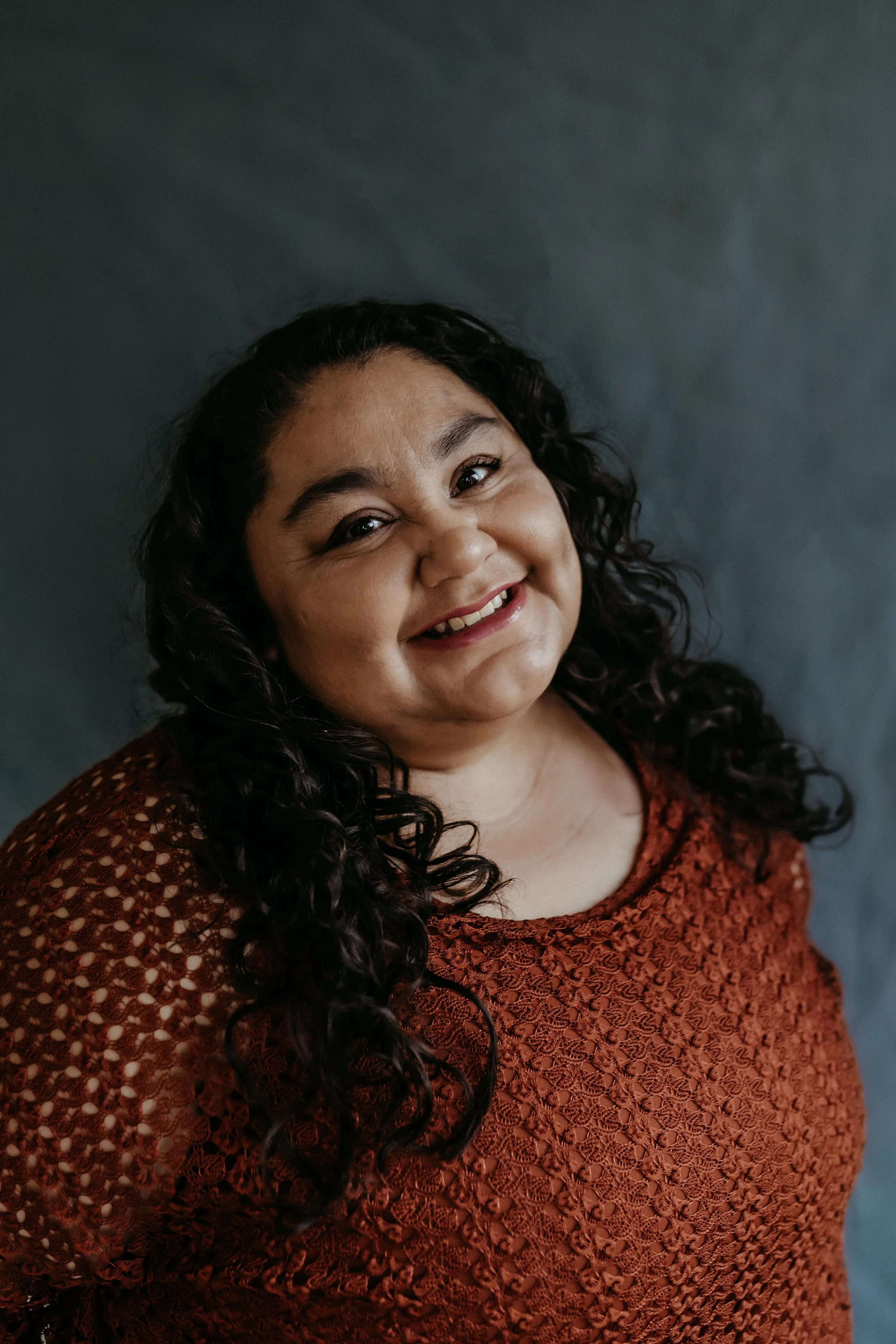 Light skinned Latina woman with curly black shoulder lengthed hair, smiling at the camera wearing a maroon colored textured blouse.