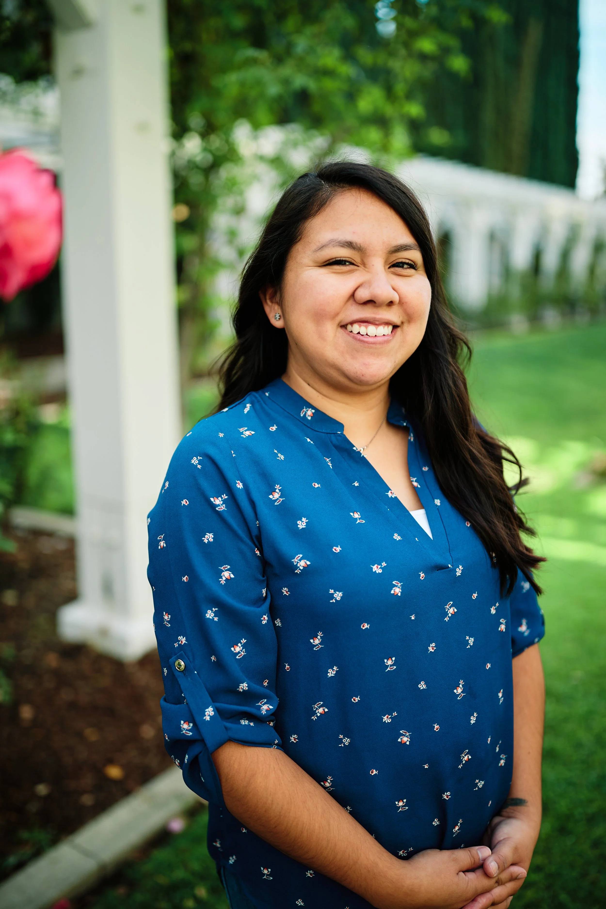 Karen, a brown-skinned Latina with long dark brown hair, in a blue floral shirt, stands smiling in a garden. Her hands are clasped in front of her, and she is smiling broadly at the camera. She also wears earrings and a necklace.