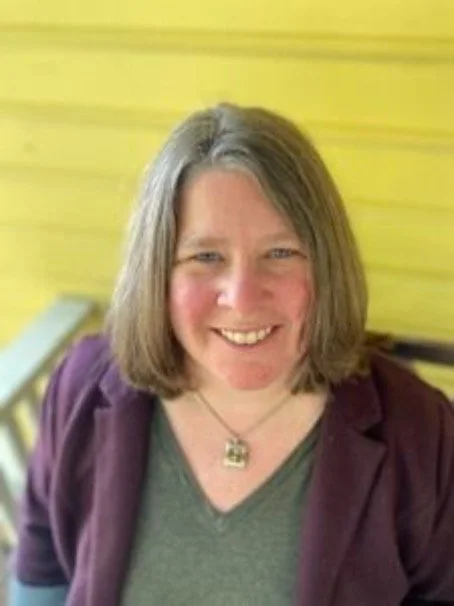 A woman with shoulder-length hair smiling, wearing a purple jacket over a green top, with a yellow wooden wall in the background.