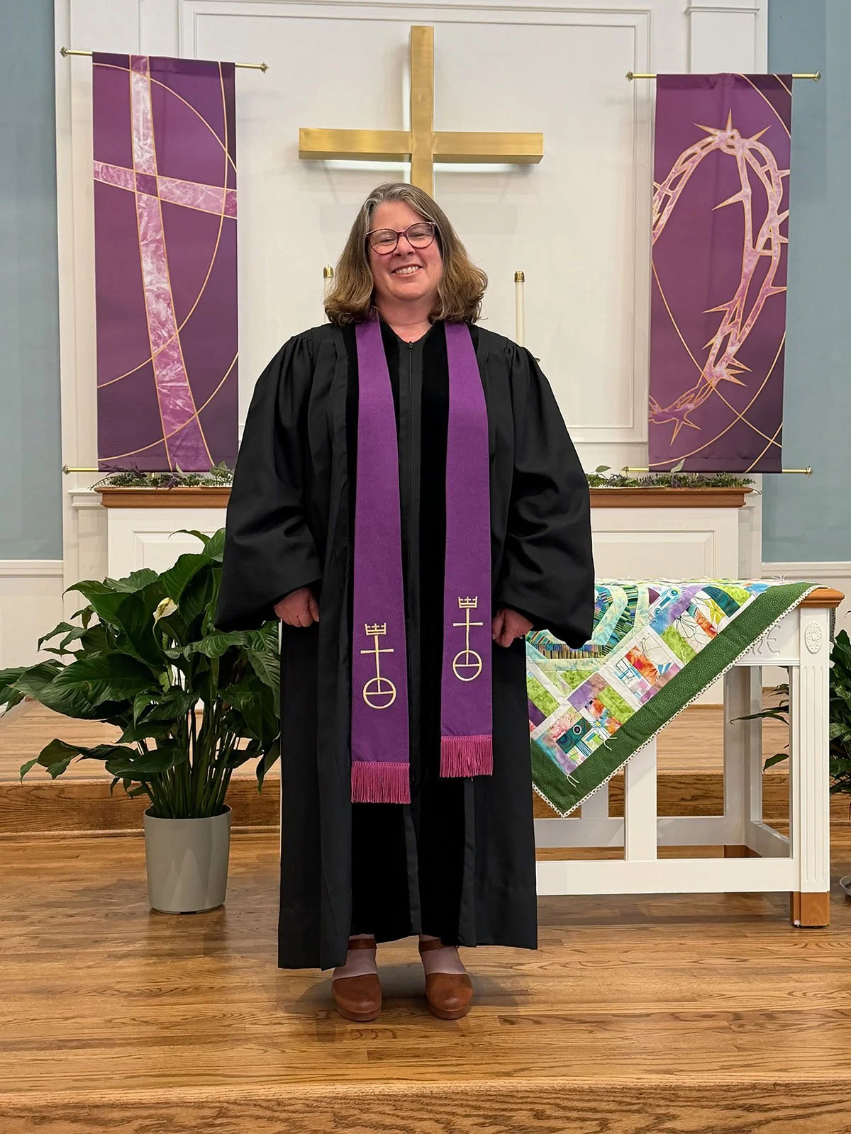 A woman with shoulder-length hair smiling, wearing a purple jacket over a green top, with a yellow wooden wall in the background.